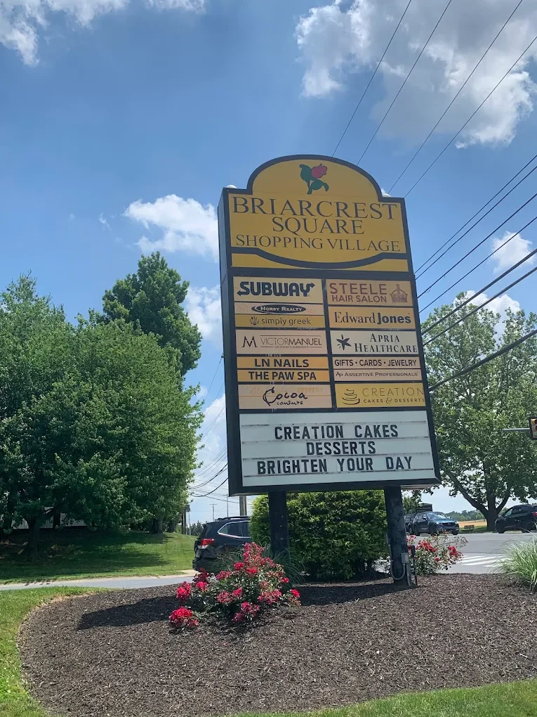 Sign for Briarcrest Square Shopping Village listing various stores and services, with a message about cakes, desserts, and brightening your day, set outdoors with trees, flowers, and a parking lot.