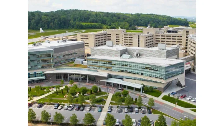 A modern hospital or medical center with a large parking lot in front, surrounded by green trees and hills in the background.