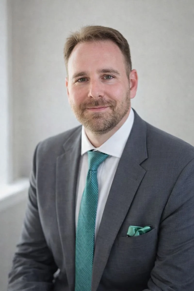 A man in a gray suit with a white shirt and teal tie, sitting against a neutral background, smiling at the camera.