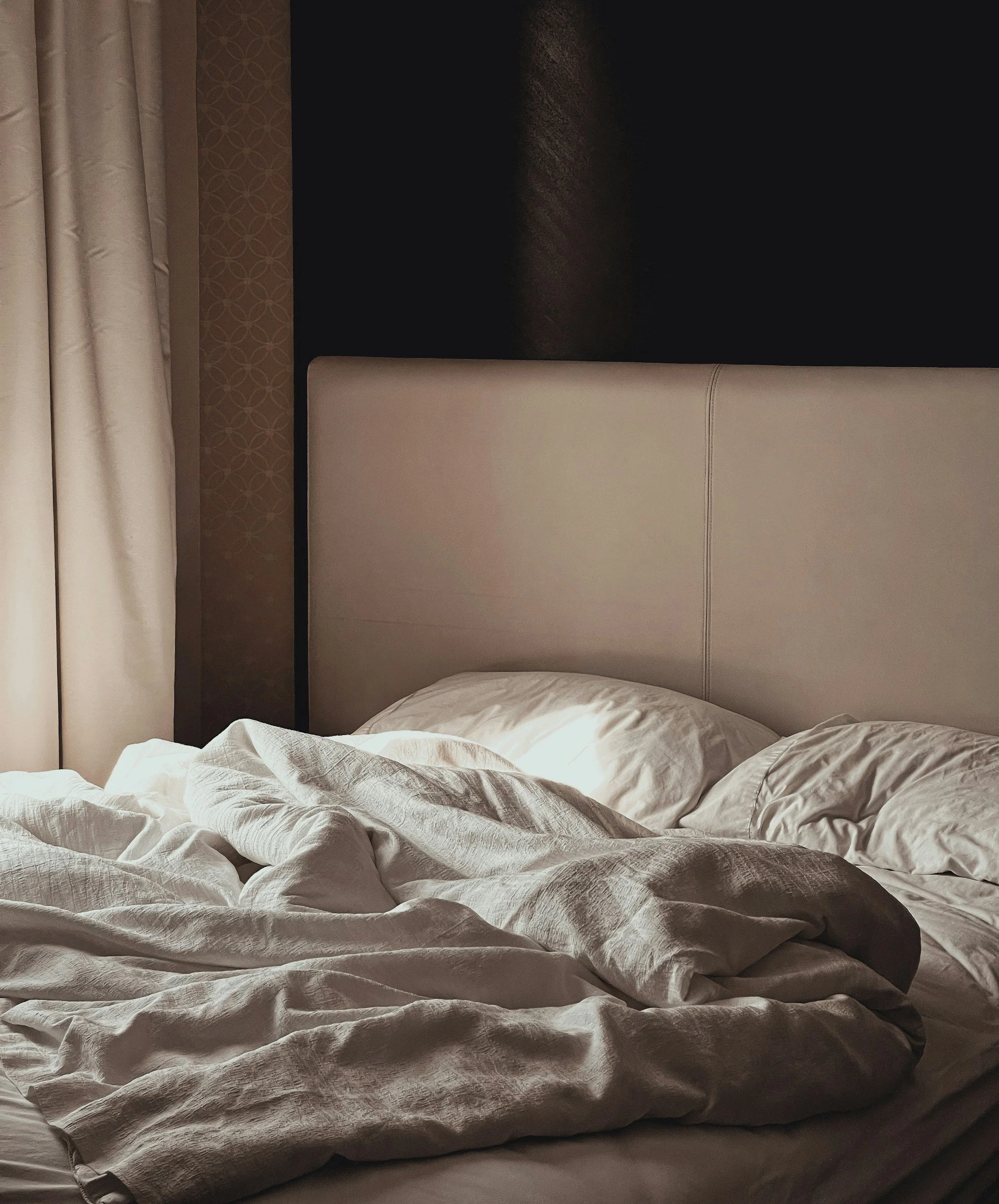 Unmade bed in a dimly lit room with a beige headboard, white pillows, and crumpled white sheets, next to a window with curtains.