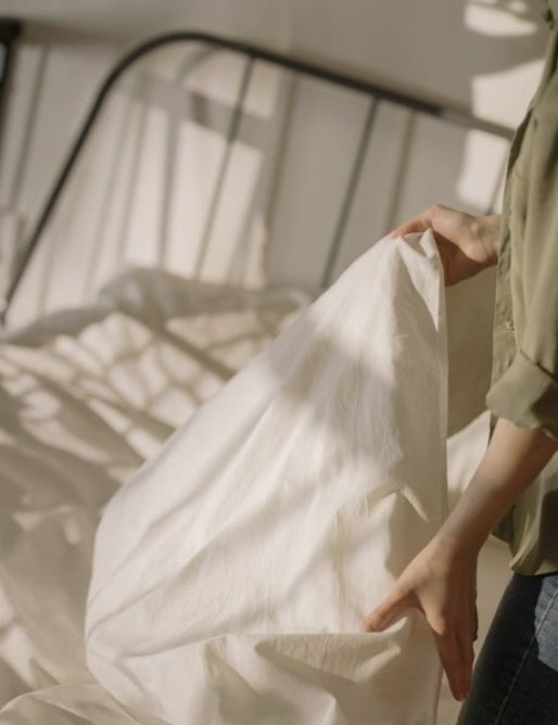 Person making the bed, pulling white sheets in a bedroom with sunlight.