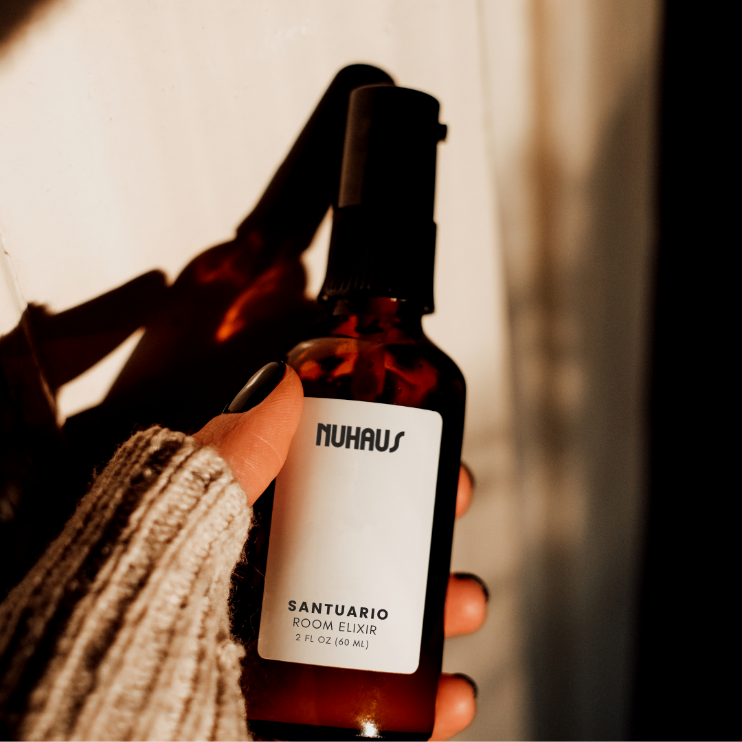 A hand holding a dark amber spray bottle labeled 'NUHAUS' and 'SANTUARIO ROOM ELIXIR' with 2 fl oz/60 ml, against a plain background with shadows.
