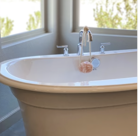 A white bathtub with two silver faucets, positioned by large windows with greenery outside, with a pink bath sponge hanging from the faucet.