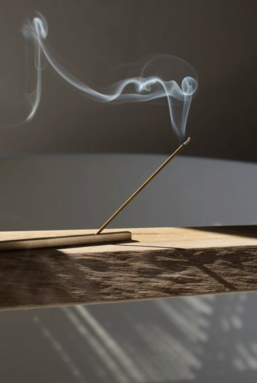 A thin incense stick burning, with smoke curling upward against a dark background and casting shadows on a wooden surface.