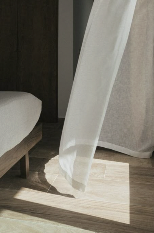 Sunlight passing through a sheer white curtain onto a wooden floor near a bed and a darker curtain.