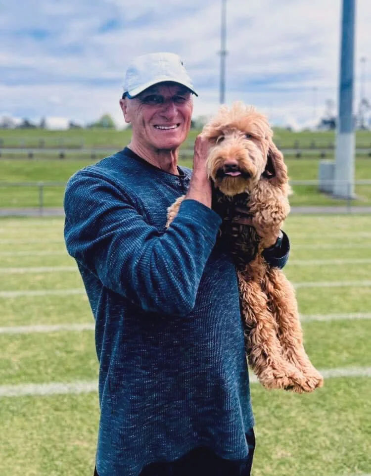 A man wearing a white cap and blue sweatshirt holding a fluffy brown dog in an outdoor setting with grassy fields and cloudy sky in the background.
