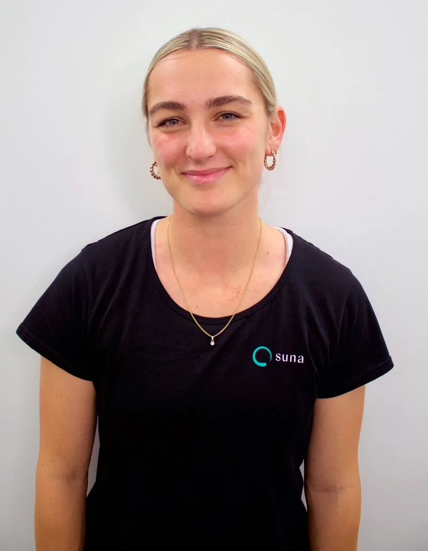 A young woman with blonde hair tied back, wearing earrings, a necklace, and a black t-shirt with the logo 'suna' on it, standing against a white background, smiling at the camera.