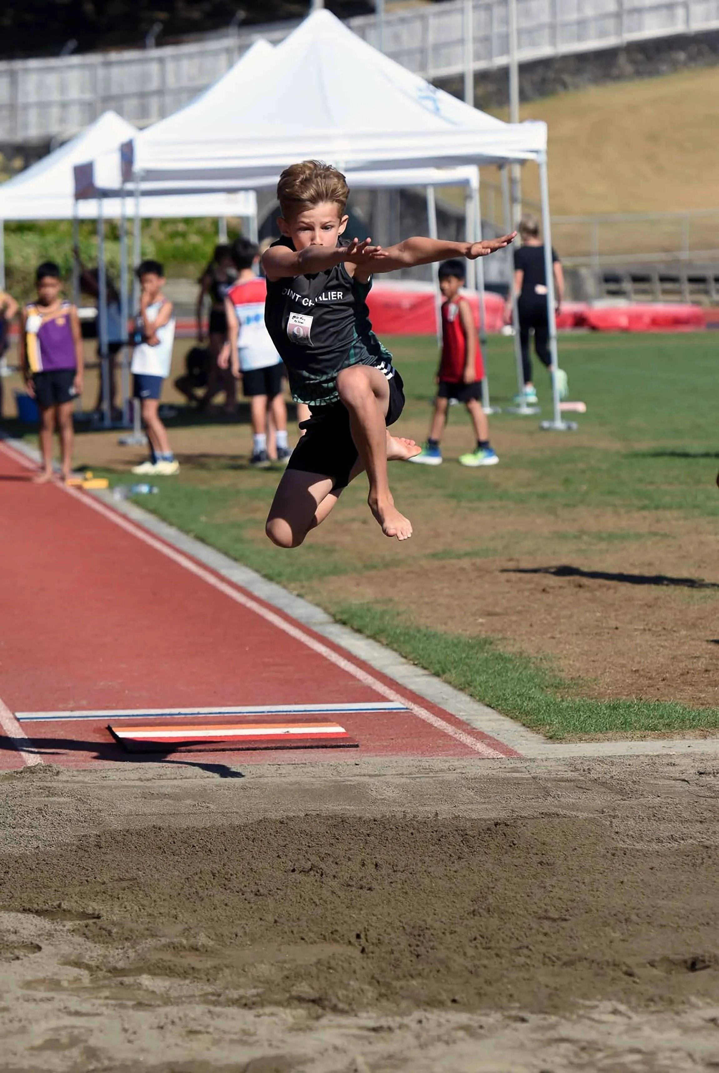 Young boy in mid-air during a long jump at a track and field event, with other children and tents in the background on a sunny day.