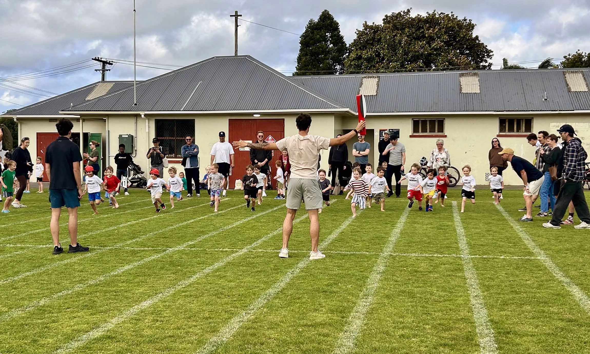 Children participating in a running race on a grassy field, with adults and spectators watching under cloudy skies.