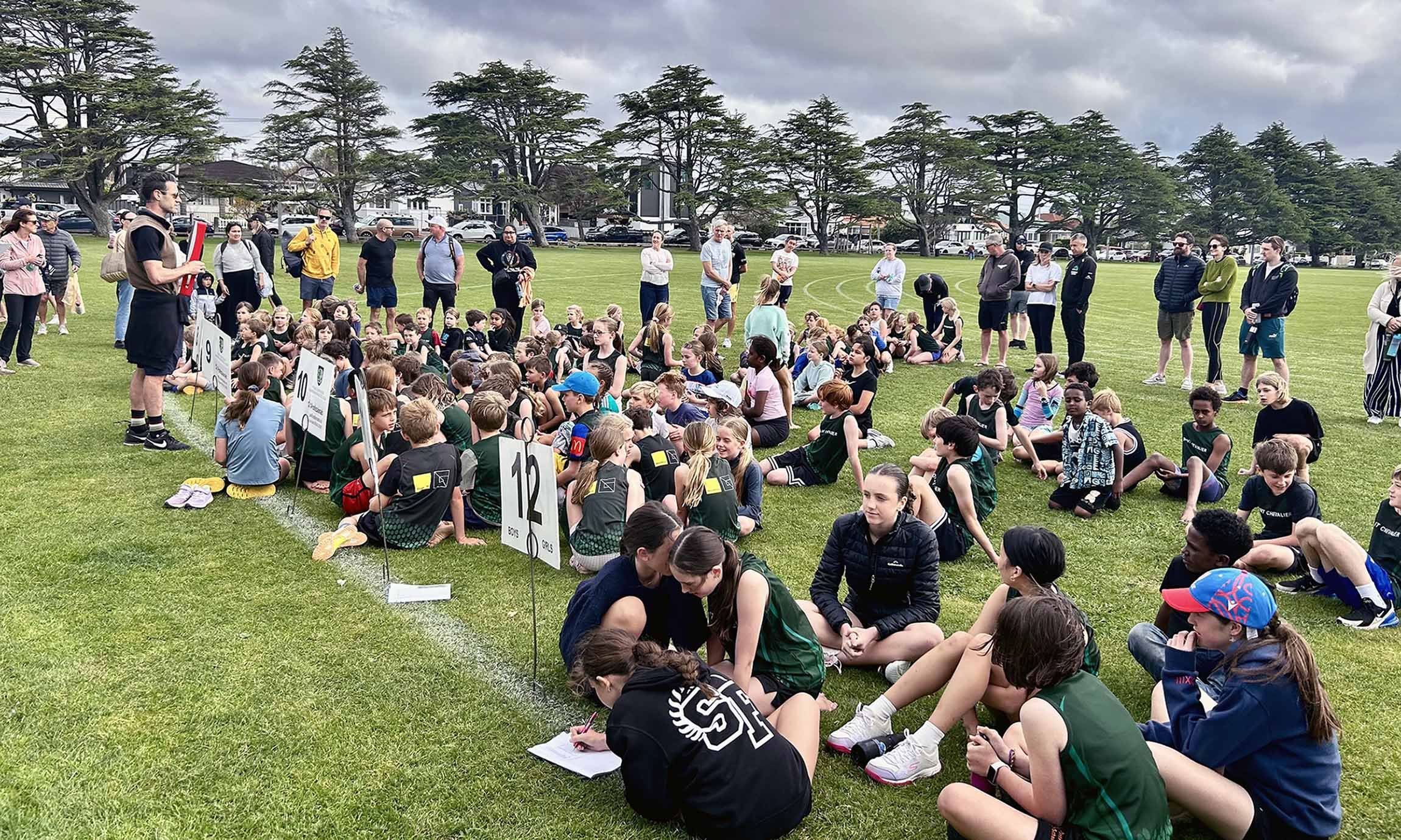 Group of students seated on grass at an outdoor sports field, listening to coach standing nearby, with parents and spectators standing behind.
