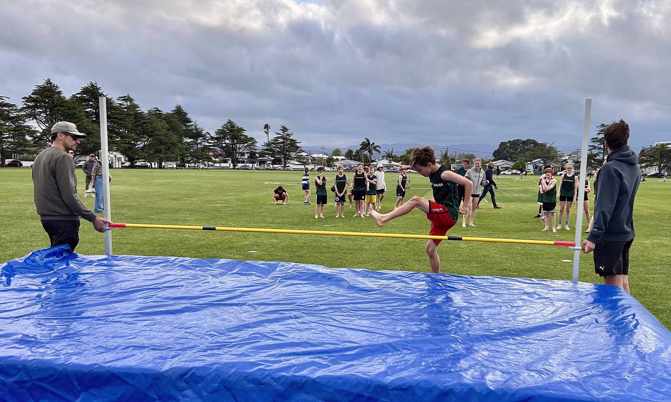 A young athlete jumps over a high jump bar at an outdoor track and field event, with onlookers watching on a grassy field under a cloudy sky.