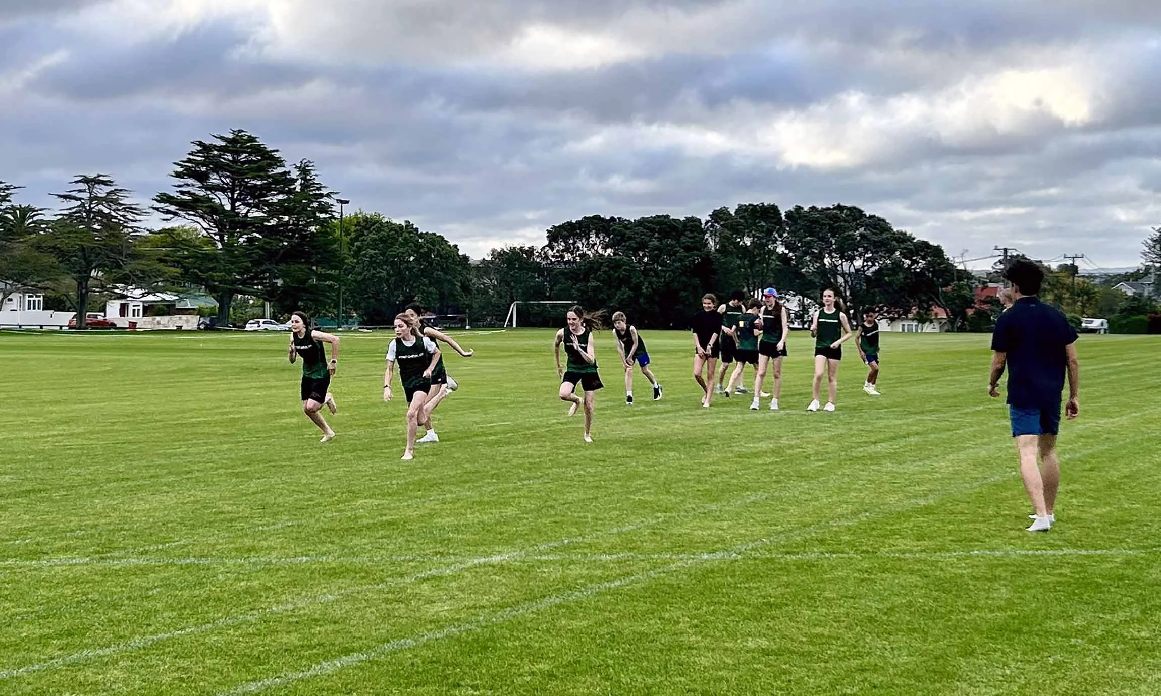 A group of young athletes running on a grassy sports field under cloudy sky, some wearing black and green sports uniforms, with trees and houses in the background.