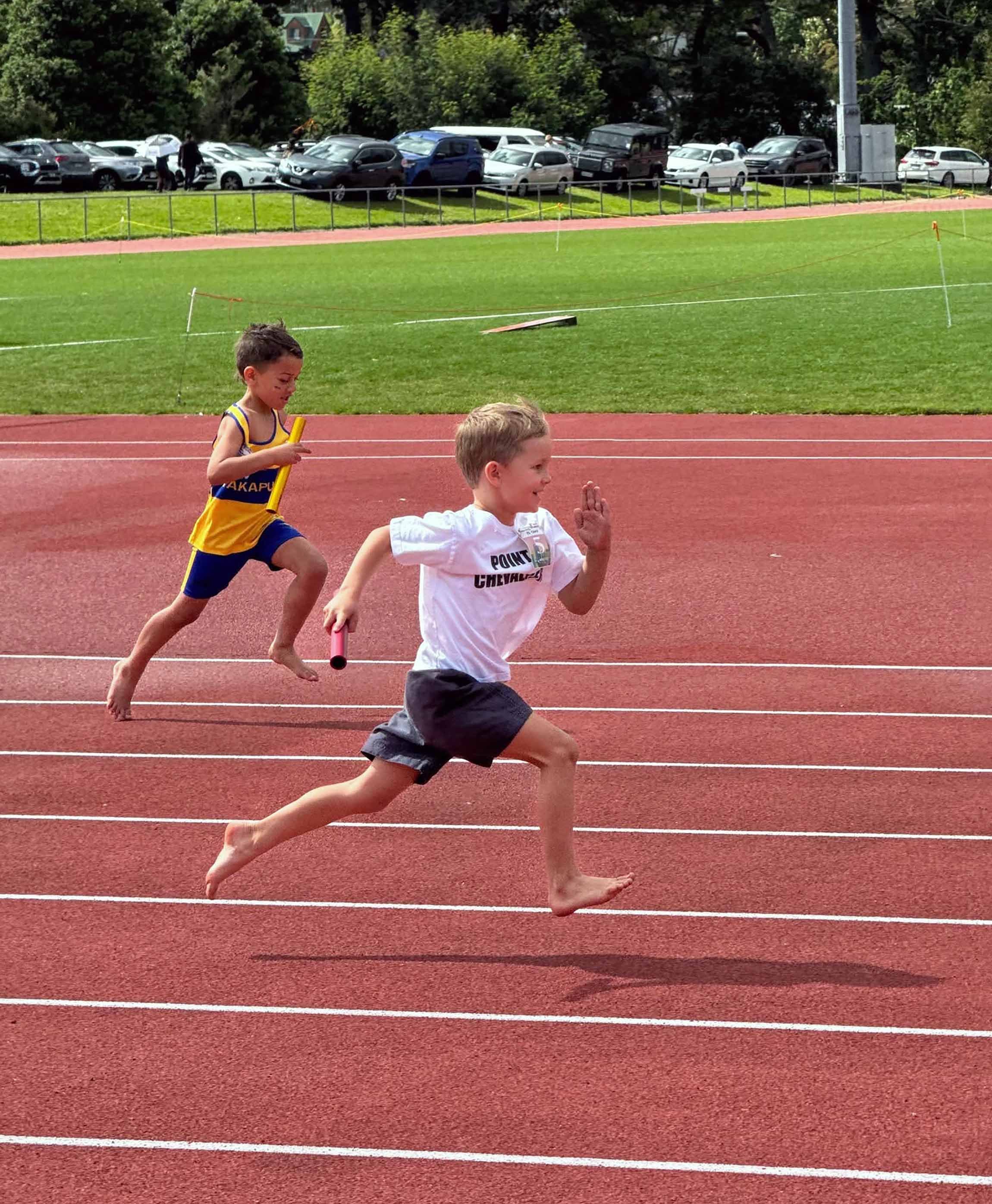 Two young boys running barefoot on a red track during a relay race, with one holding a baton. One boy in front is wearing a white T-shirt and dark shorts, while the other behind wears a yellow and blue athletic outfit. Cars and greenery are visible in the background.