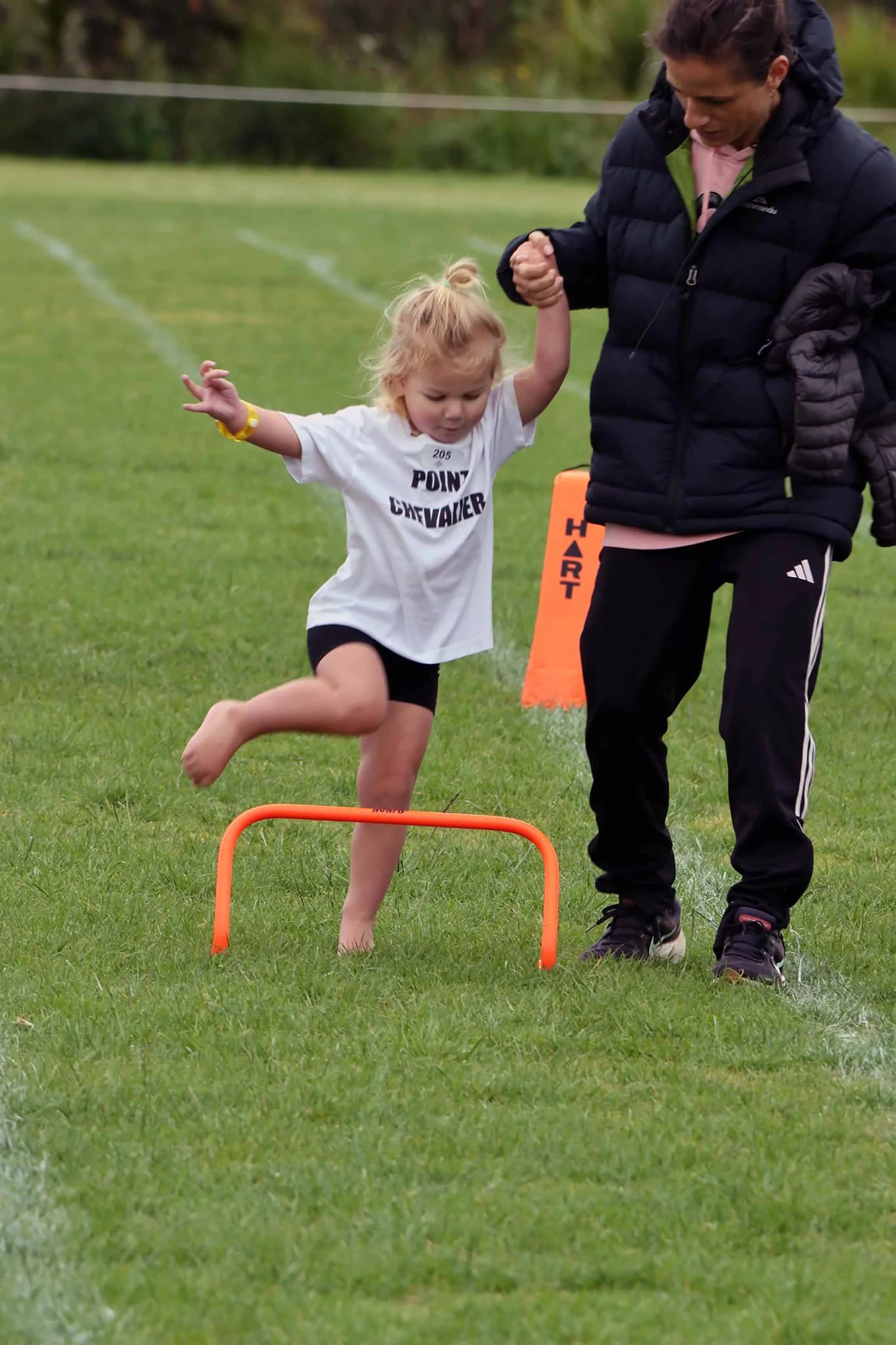 A young girl running while jumping over a small orange hurdle on a grassy field during an athletic event, assisted by an adult woman.