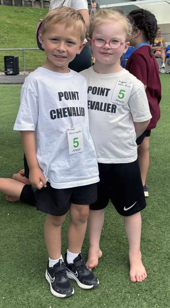 Two young children, a boy and a girl, standing on a grassy field at an outdoor event. They are wearing white T-shirts with black text that reads "POINT CHEVALIER" and have green "5" stickers on their shirts. The boy is wearing black shorts and black sneakers, while the girl is barefoot and has glasses with pink frames. Other children and people are visible in the background.