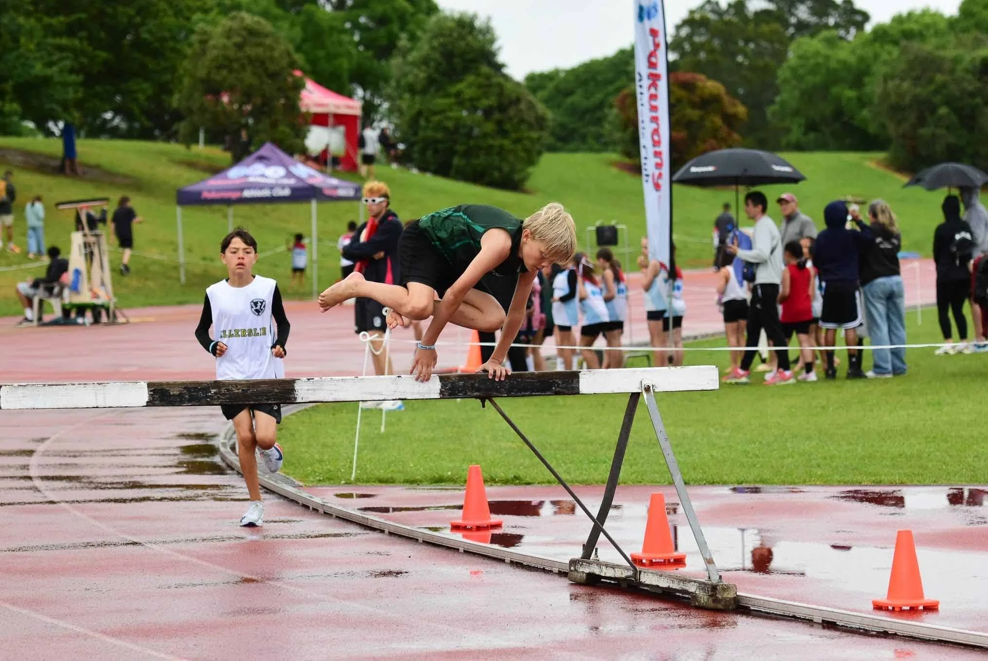 A young male athlete is attempting to jump over a high bar during a track and field event, while another young athlete runs towards the bar on a wet track. Several spectators and athletes are gathered in the background, some holding umbrellas, on a green outdoor sports field on a rainy day.