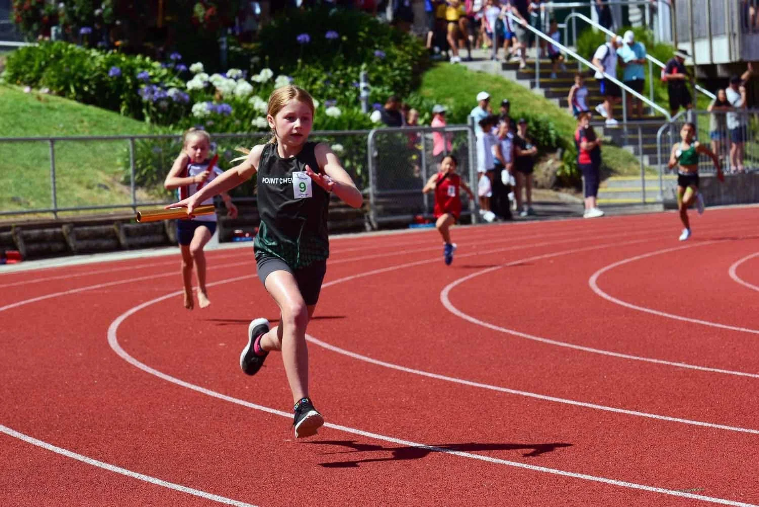 Children running on a red track during a race at an outdoor stadium, with spectators watching in the background.