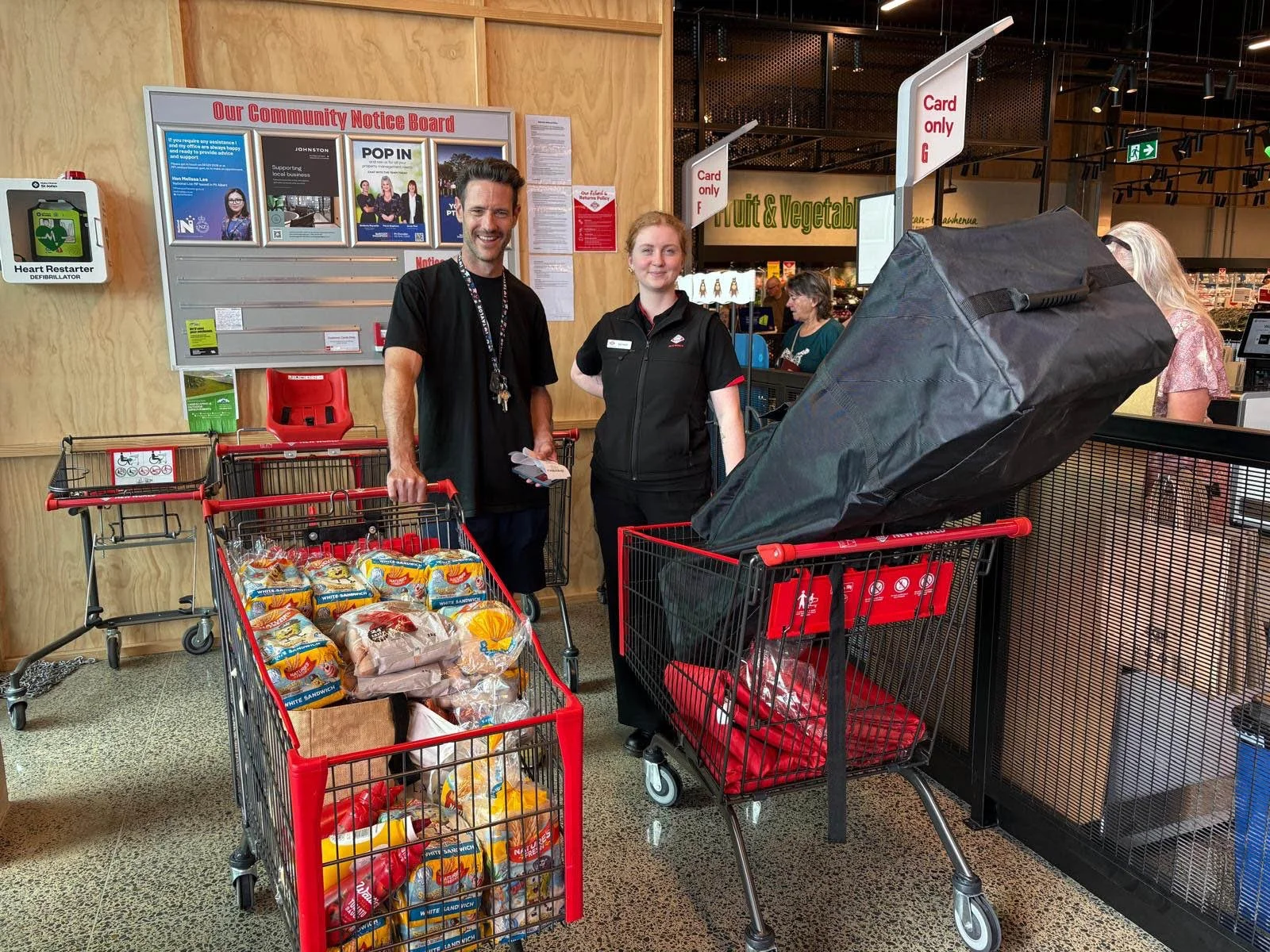 A man and a woman standing at a checkout counter in a grocery store, with shopping carts filled with bread and large black bag, in front of a community notice board.