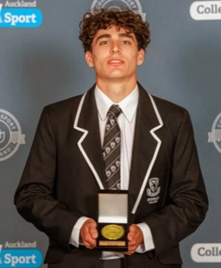 A young man in a black blazer with white piping, white shirt, and striped tie is holding a gold medal in a box. He is standing in front of a backdrop with logos and text, likely at an award or recognition event.