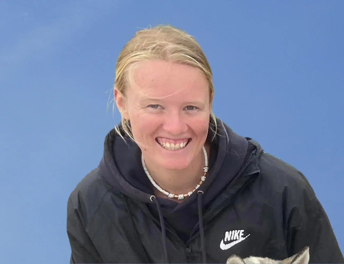 Smiling woman with blonde hair tied back, wearing a black Nike hoodie and a shell necklace, in front of a blue background.