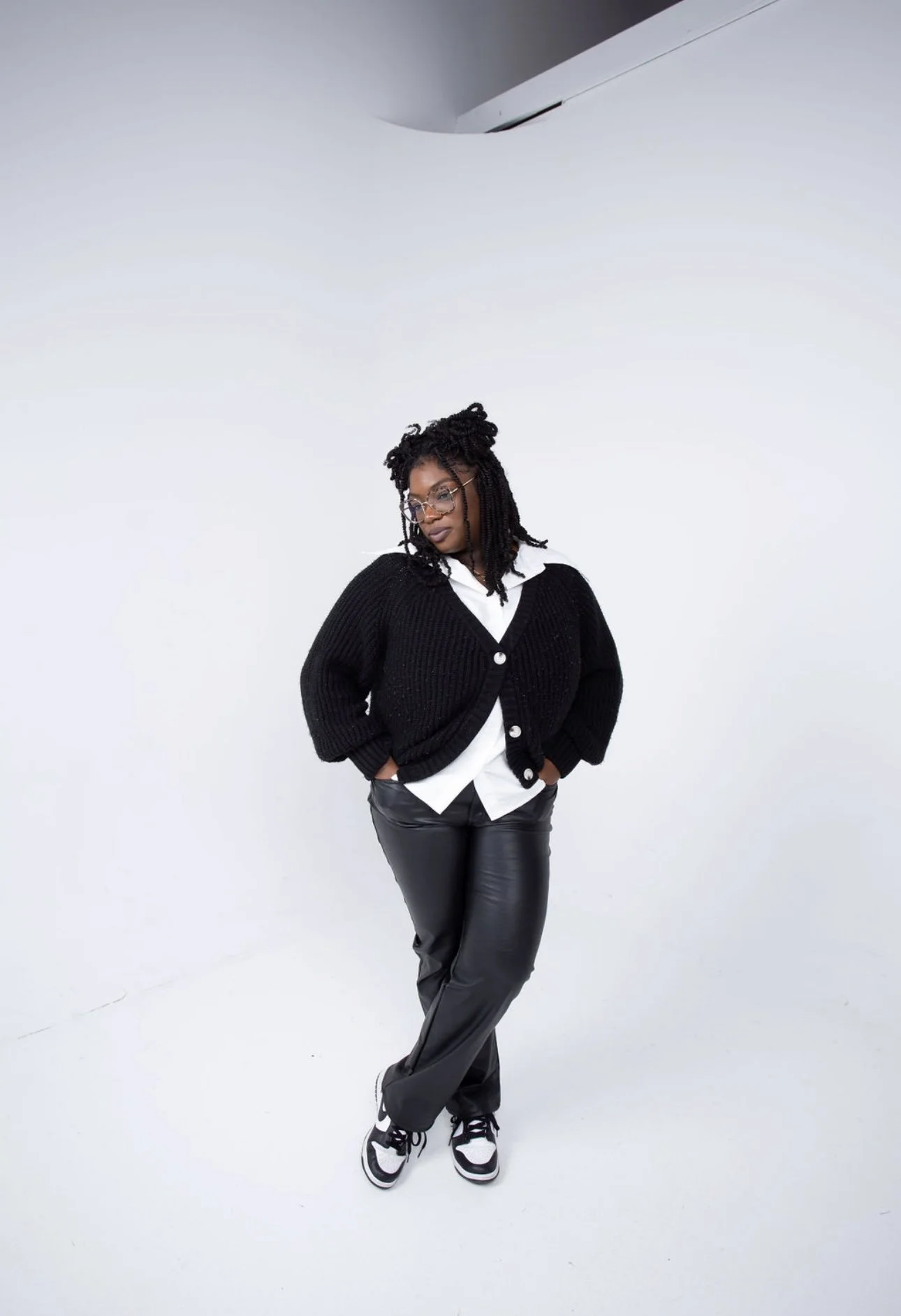A woman standing against a white background, dressed in a black sweater, white shirt, black leather pants, and sneakers, with her hands in her pockets and looking down.