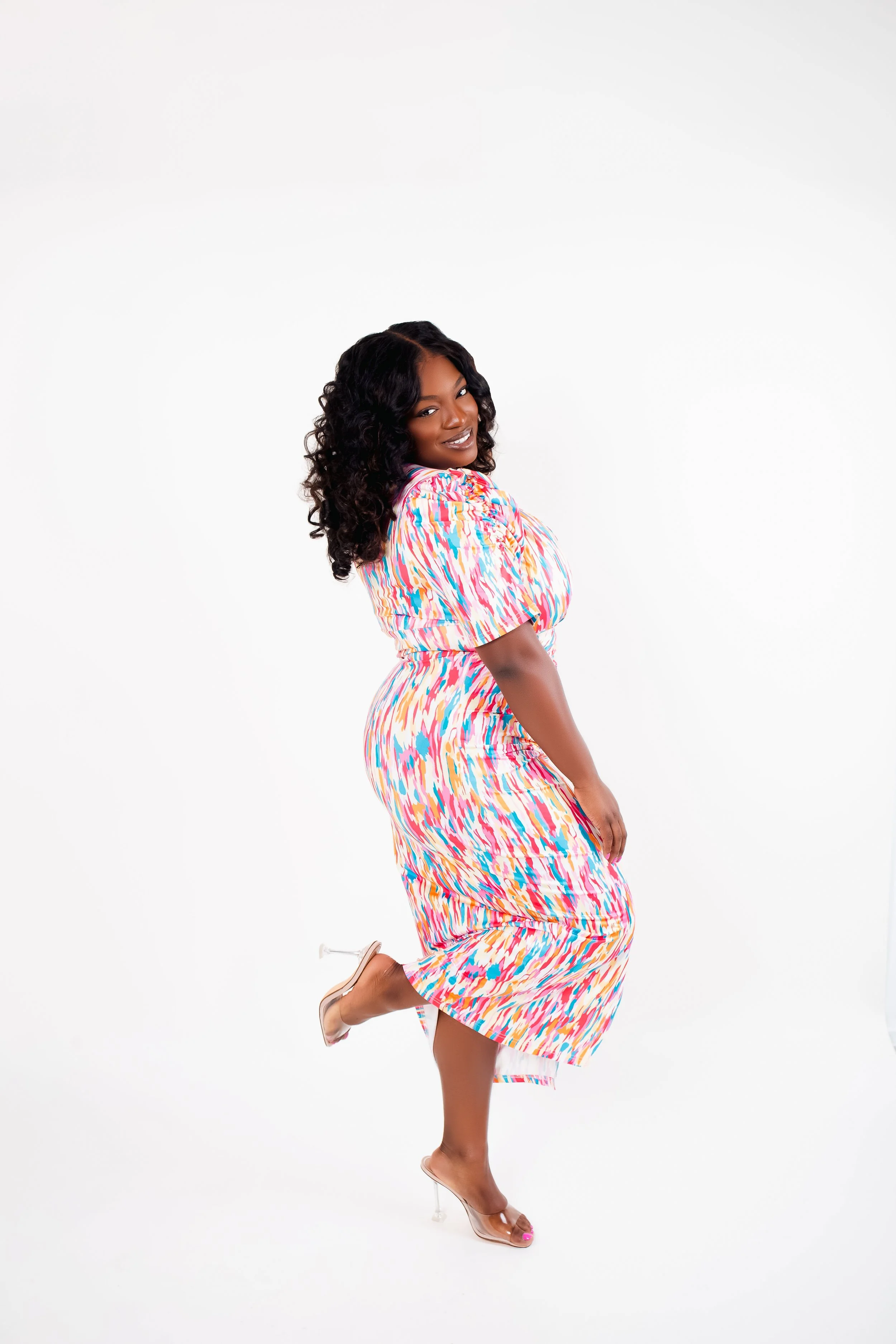 A woman with dark curly hair wearing a colorful, patterned dress and clear high heels, posing against a plain white background.