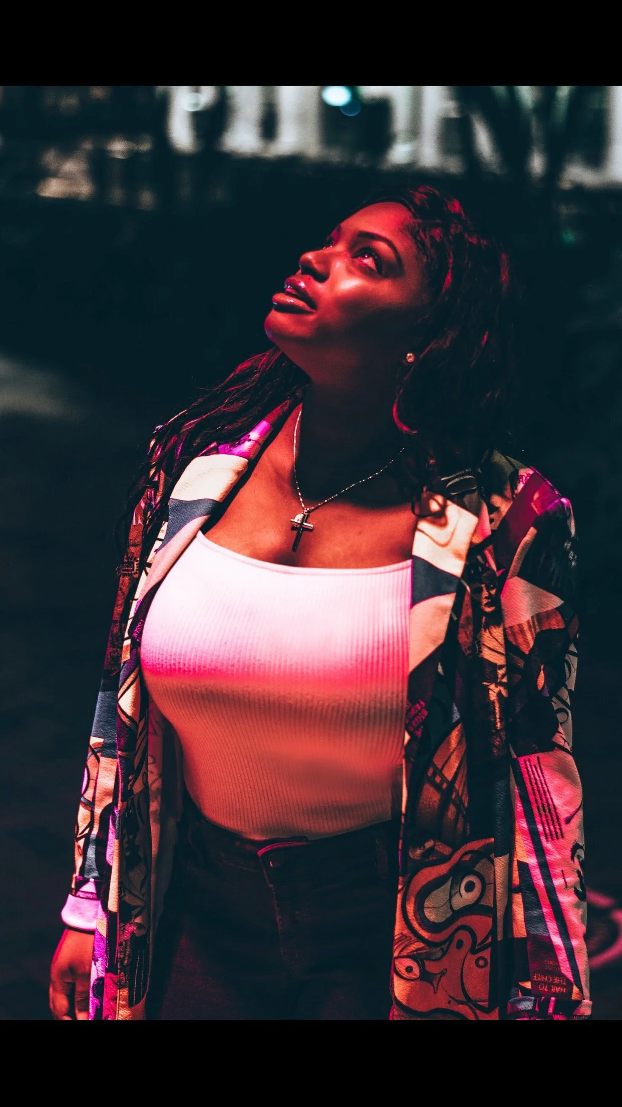 A woman with dark hair wearing a white tank top, a colorful patterned jacket, and a cross necklace, looking upwards at night.