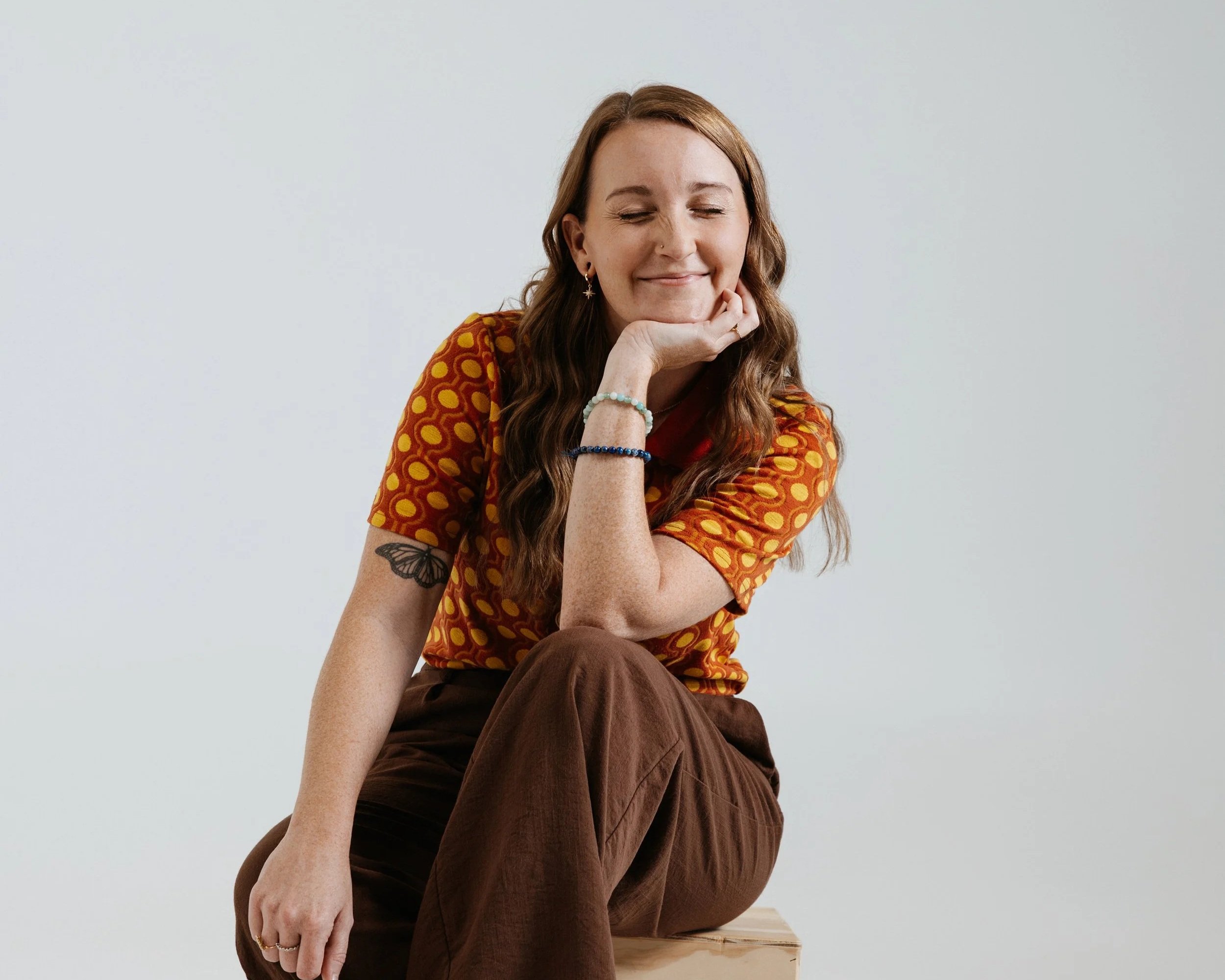 A young woman with long, wavy red hair, closed eyes, and a peaceful smile, sitting on a wooden surface against a plain, light-colored background. She rests her chin on her hand, wearing a multicolored patterned shirt, multiple bracelets, earrings, and a ring, with a butterfly tattoo on her arm.