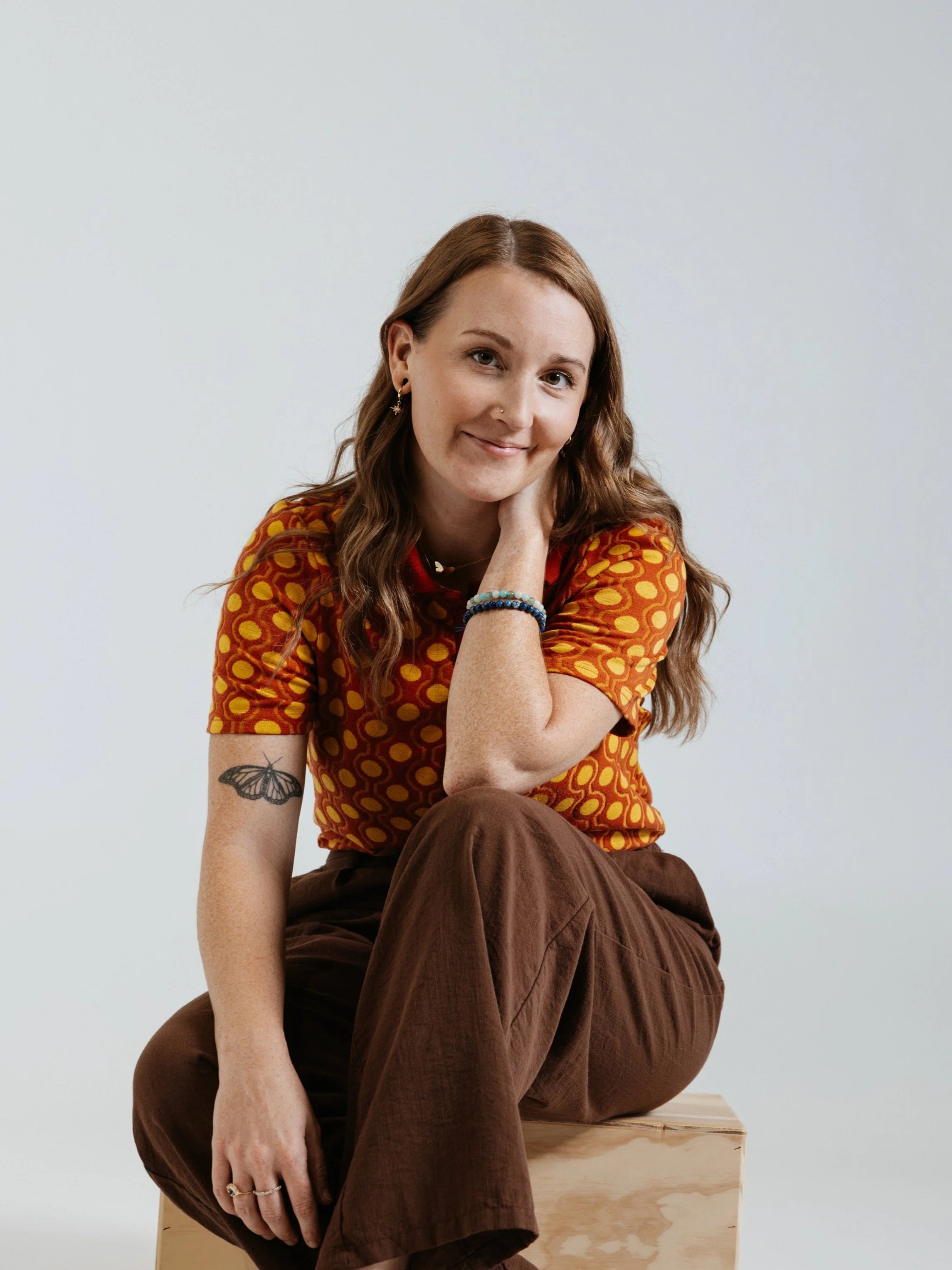 A young woman with long brown hair, wearing a patterned orange and yellow shirt, sits on a wooden block, smiling gently at the camera.