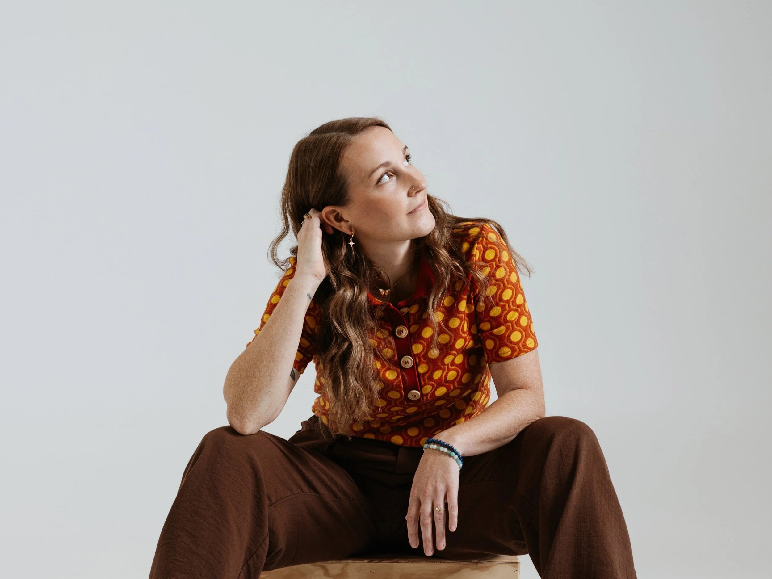 A woman with long, wavy brown hair, wearing a colorful patterned shirt and brown pants, sits with her hand resting on her head, looking to the side against a plain light gray background.