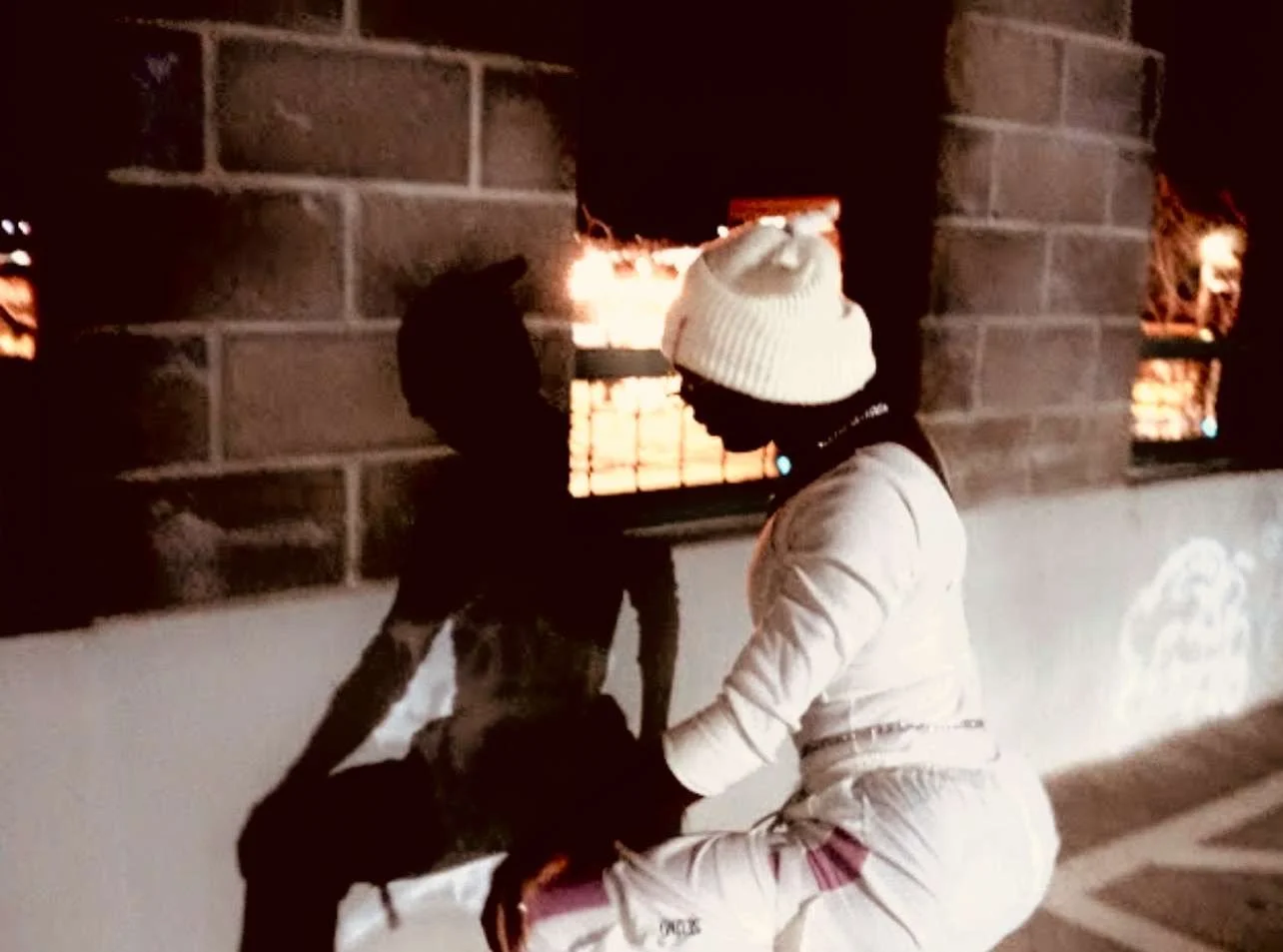 Two children in winter clothing beside a lit brick oven, one child in a white knit hat and beige coat, and the other in dark clothing, during winter evening.