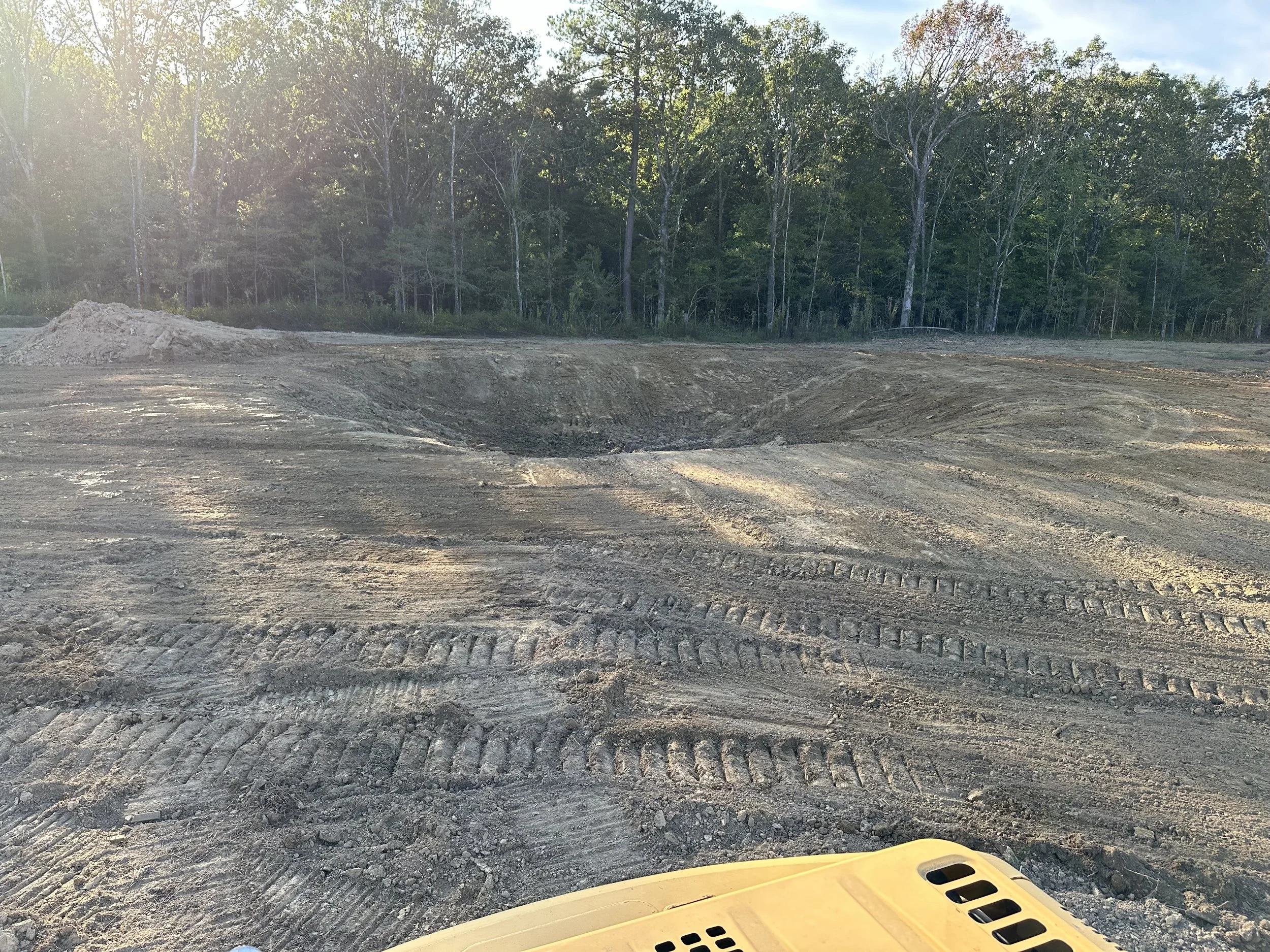 Construction site with a large hole in the ground, surrounded by dirt and tire tracks, and a forested area in the background.