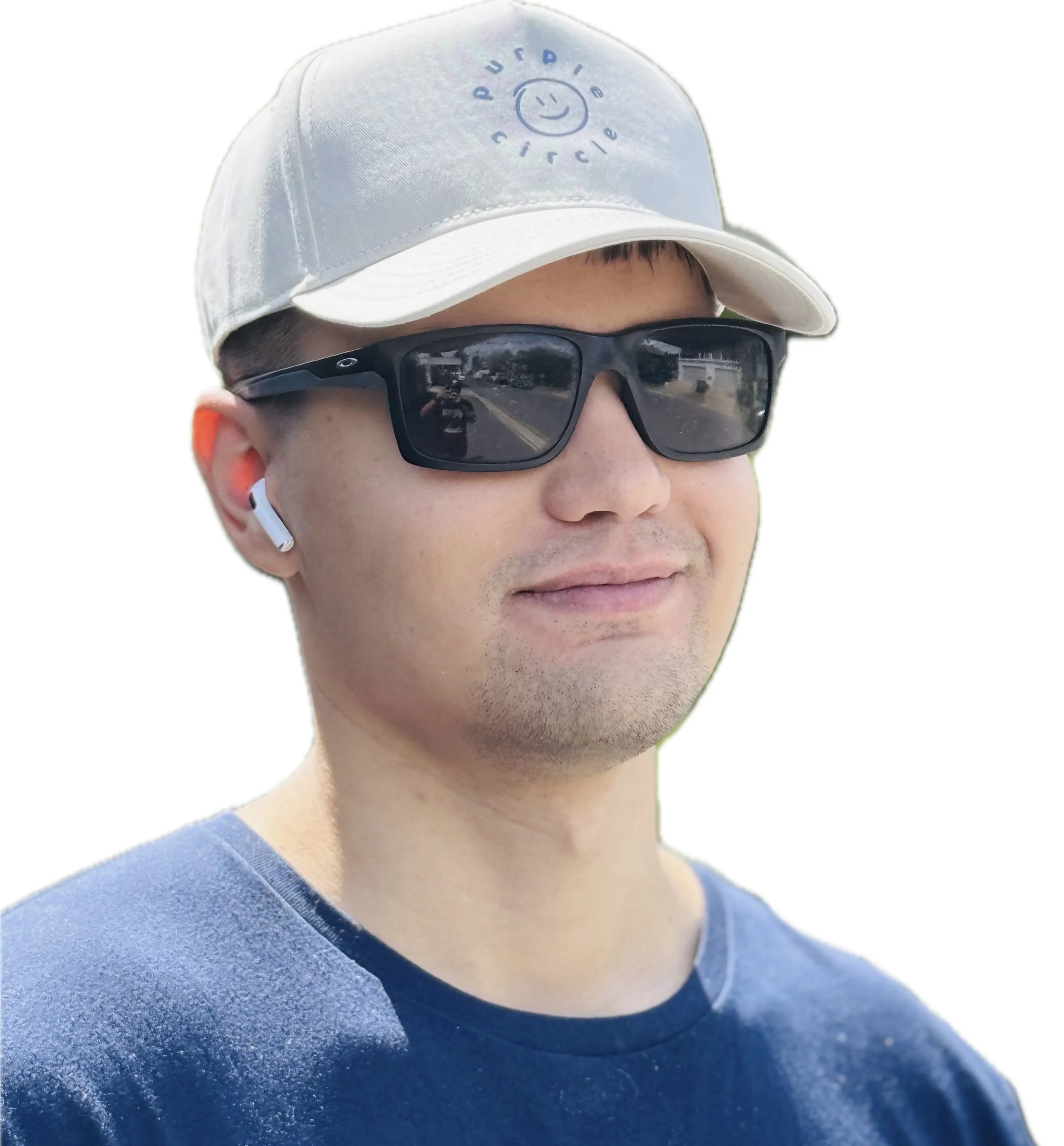 A man wearing a white cap with a smiley face and the phrase ‘purple circle,’ black sunglasses, white wireless earbuds, and a navy blue shirt, on a bright outdoor day.