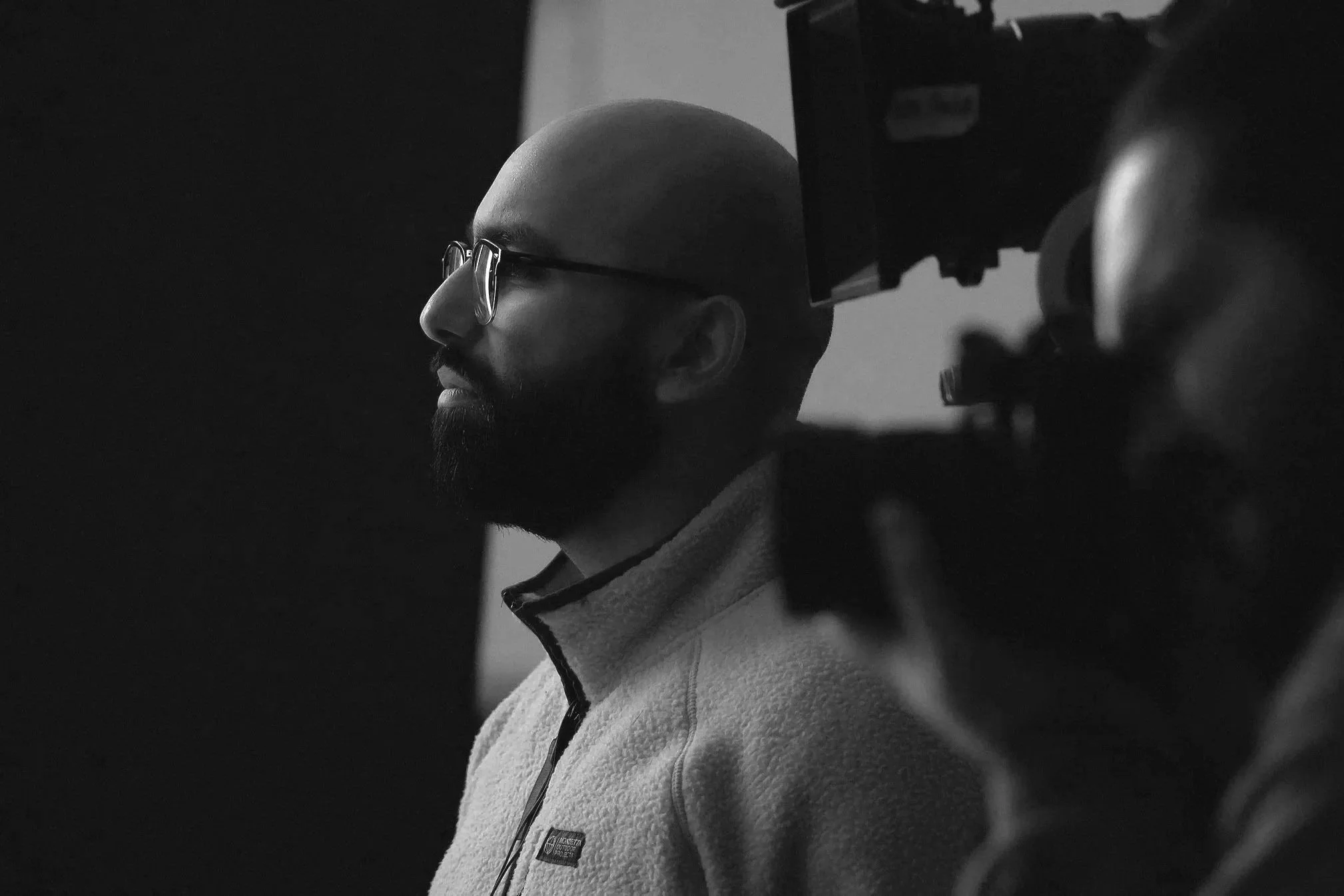 A black-and-white photo of a man with glasses and a beard, wearing a fleece jacket, looking to the left. The background is dark and out of focus.