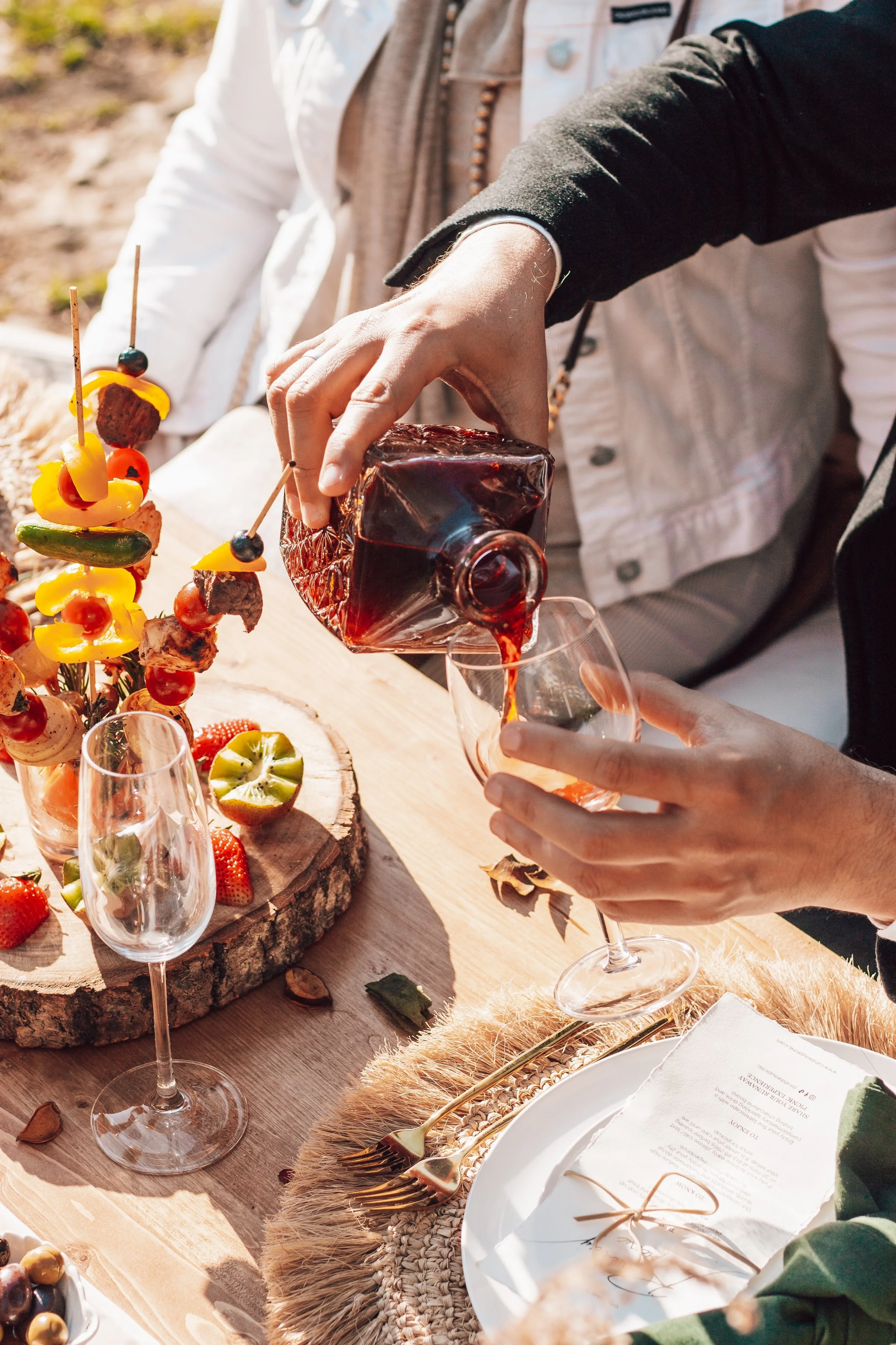 People serving red wine at an outdoor dining table decorated with fruit skewers, a wooden slice platter with strawberries and kiwis, and a place setting with gold utensils and a white napkin.