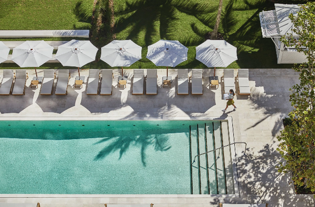 Poolside area with lounge chairs and umbrellas, green grassy area with trees, and a woman walking on the sidewalk.