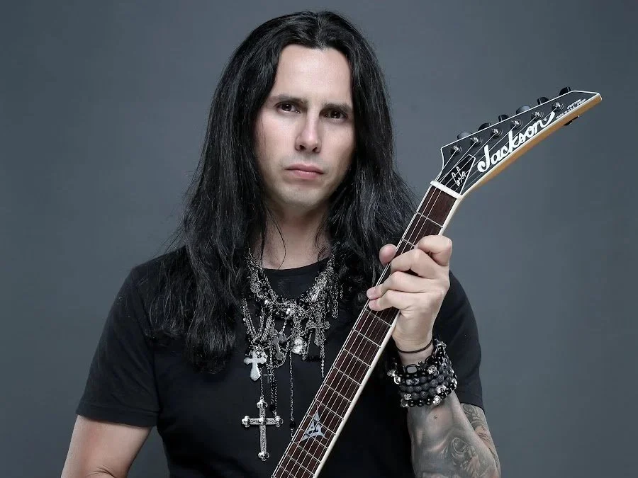 A man with long black hair, wearing a black shirt, holding an electric guitar against a gray background