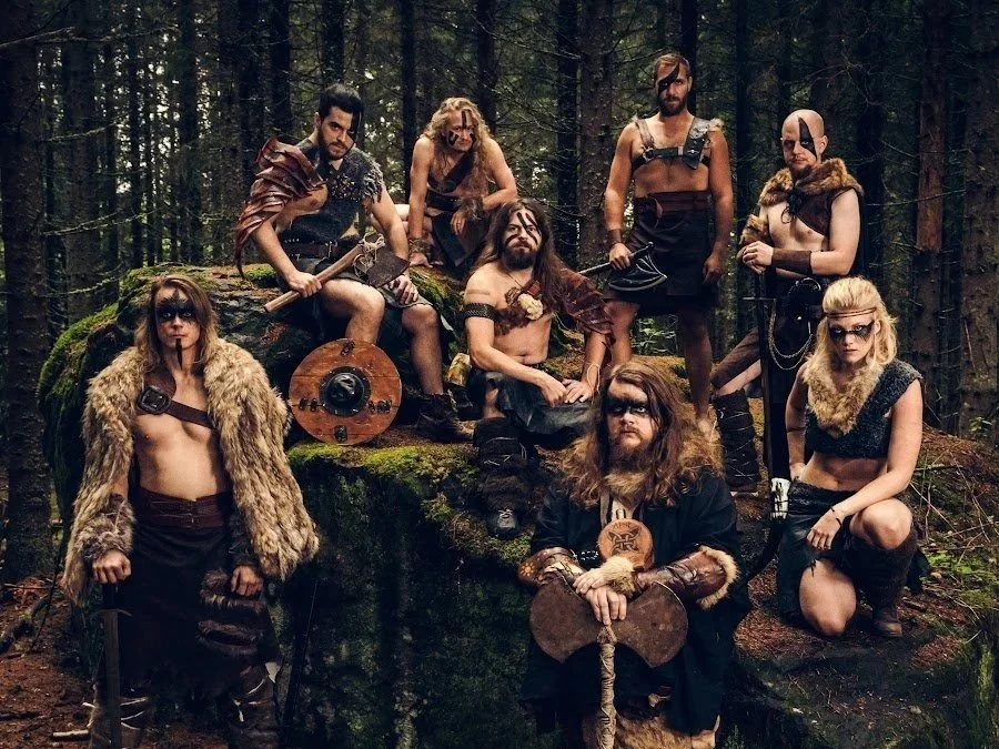 Group of eight people dressed as warriors or barbarians in a forest setting, with some holding weapons like axes and shields, and others wearing fur and leather clothing, posing on and around a moss-covered rock.