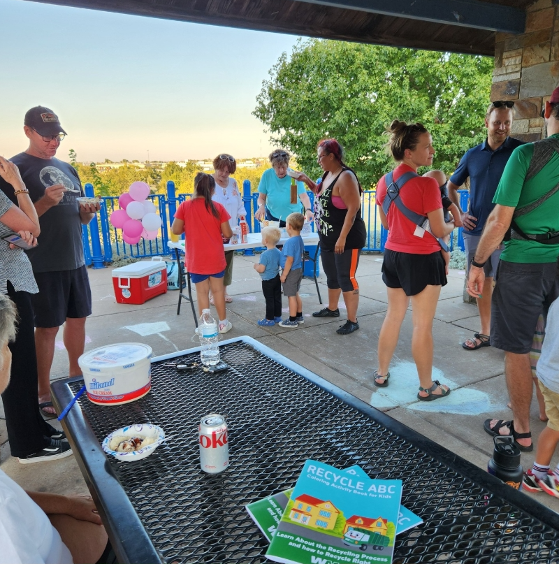 Group of adults and children gathered outside under a roof, with balloons and a table for an outdoor event, in a park or playground setting.