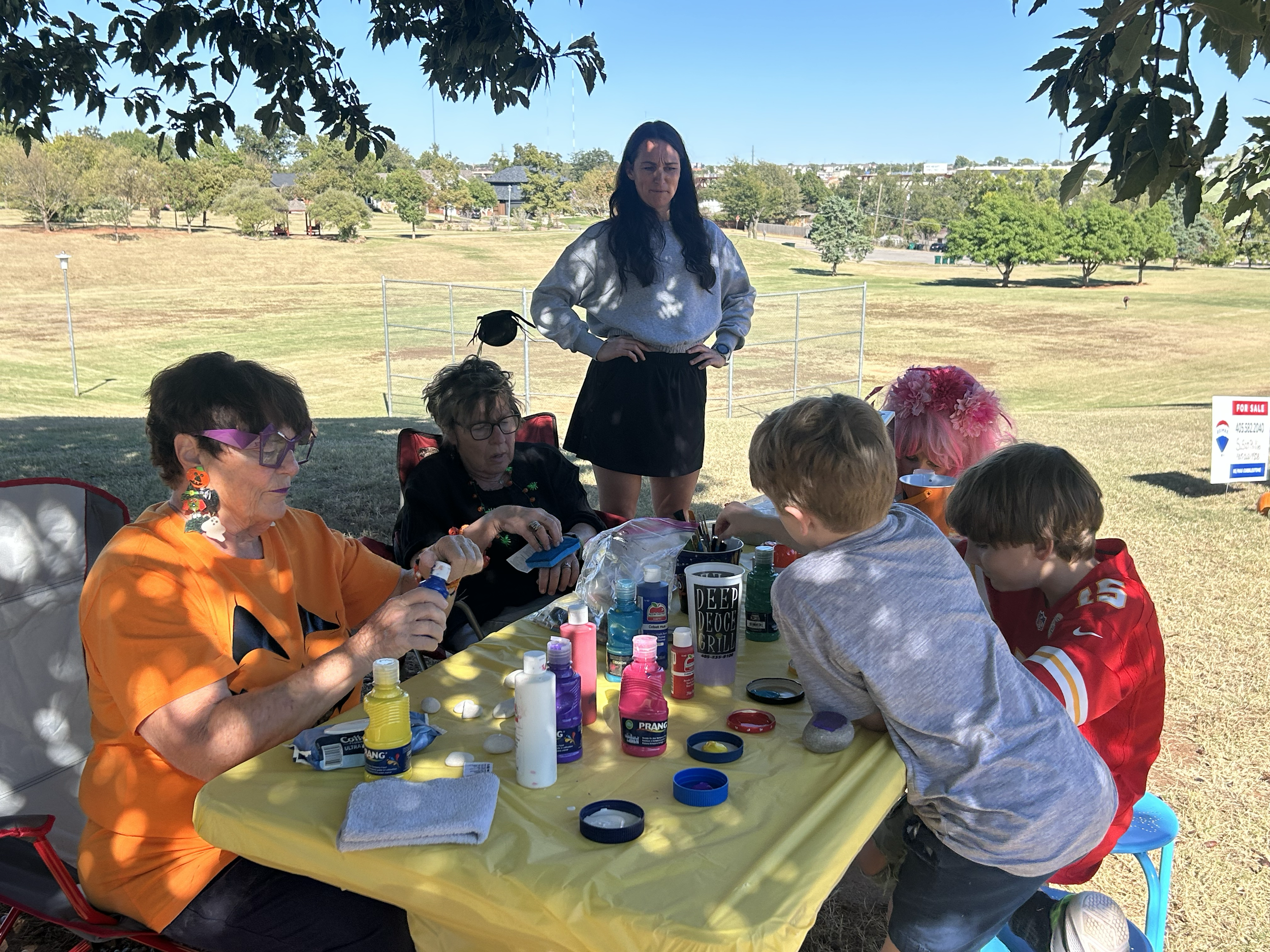 People gathered around a table outside under a tree, engaging in arts and crafts activities, with a woman standing behind them, in a grassy park.