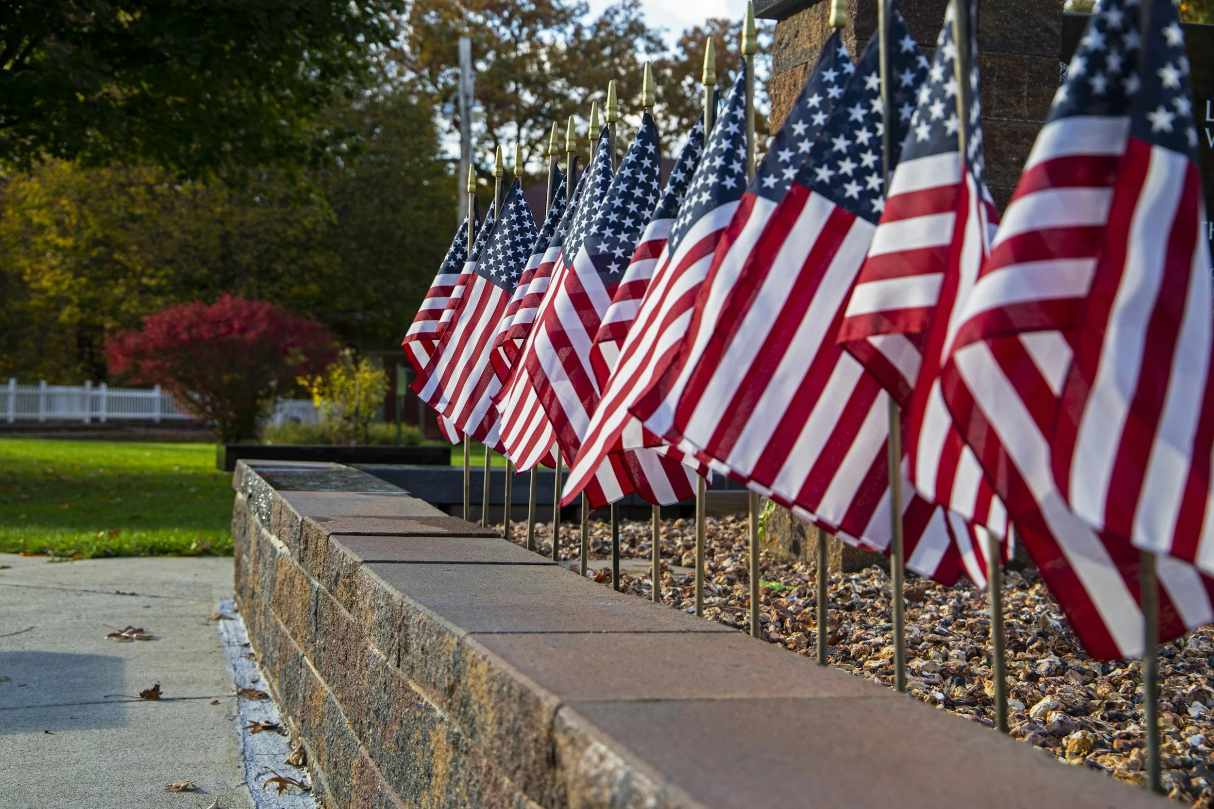 A row of American flags displayed along a brick wall outdoors, with trees and greenery in the background.