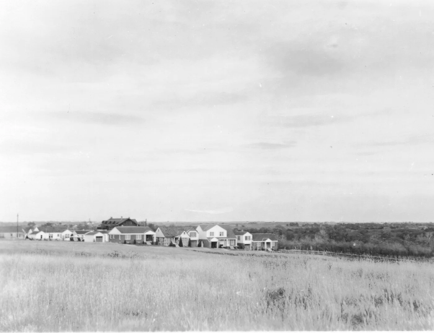 A black and white photo of a rural landscape with a field of grass in the foreground, a row of houses in the middle ground, and a cloudy sky above.