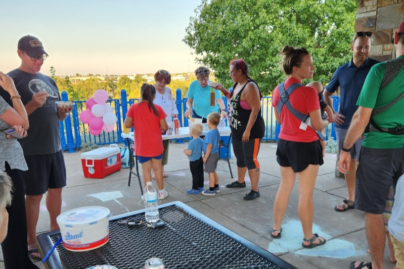 Group of people, including children and adults, gathered outdoors on a terrace with a blue railing, participating in a social event with balloons, tables, and refreshments.