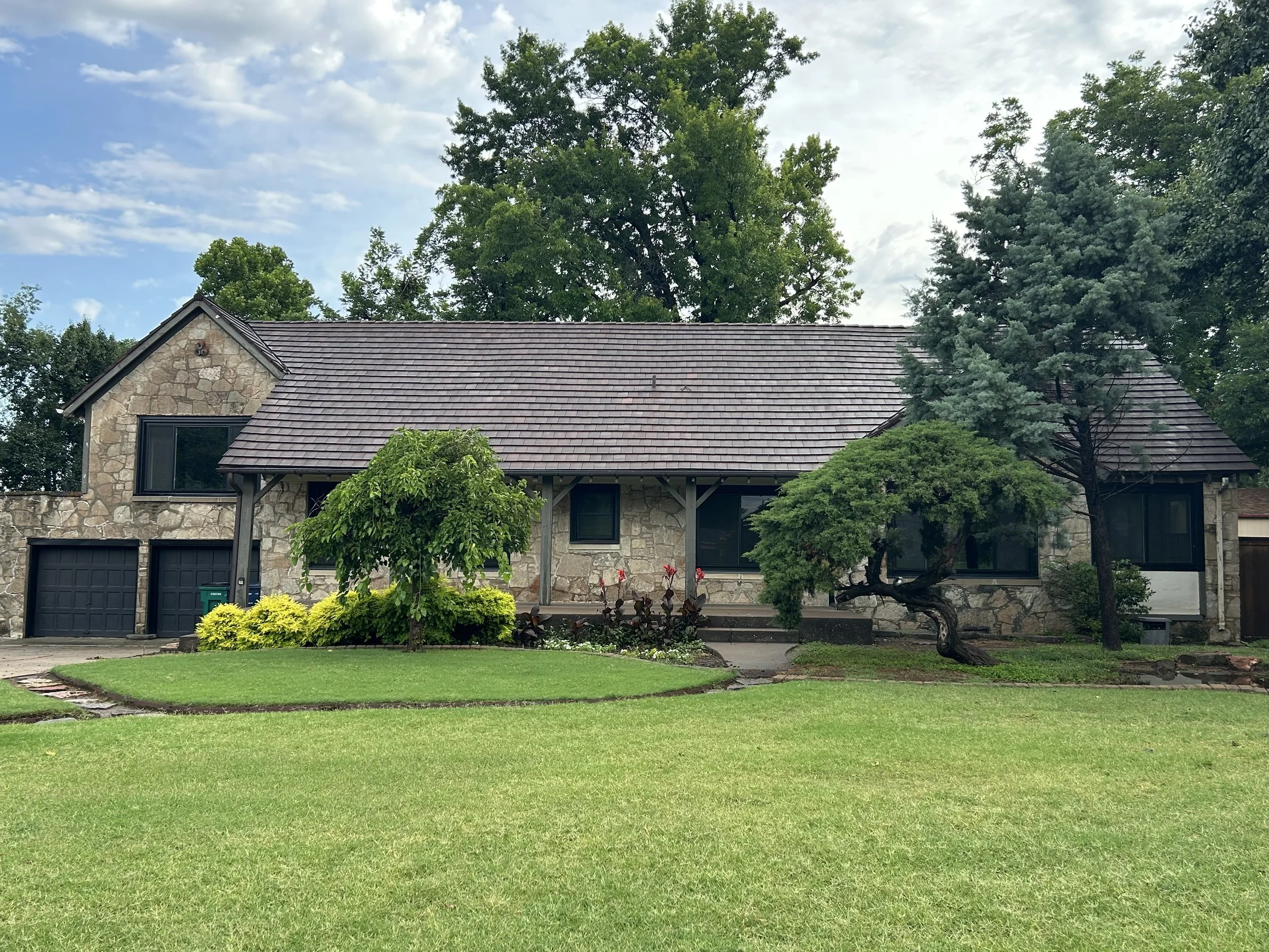 A stone house with a dark shingle roof, black window frames, and a garage, surrounded by green lawns and trees.