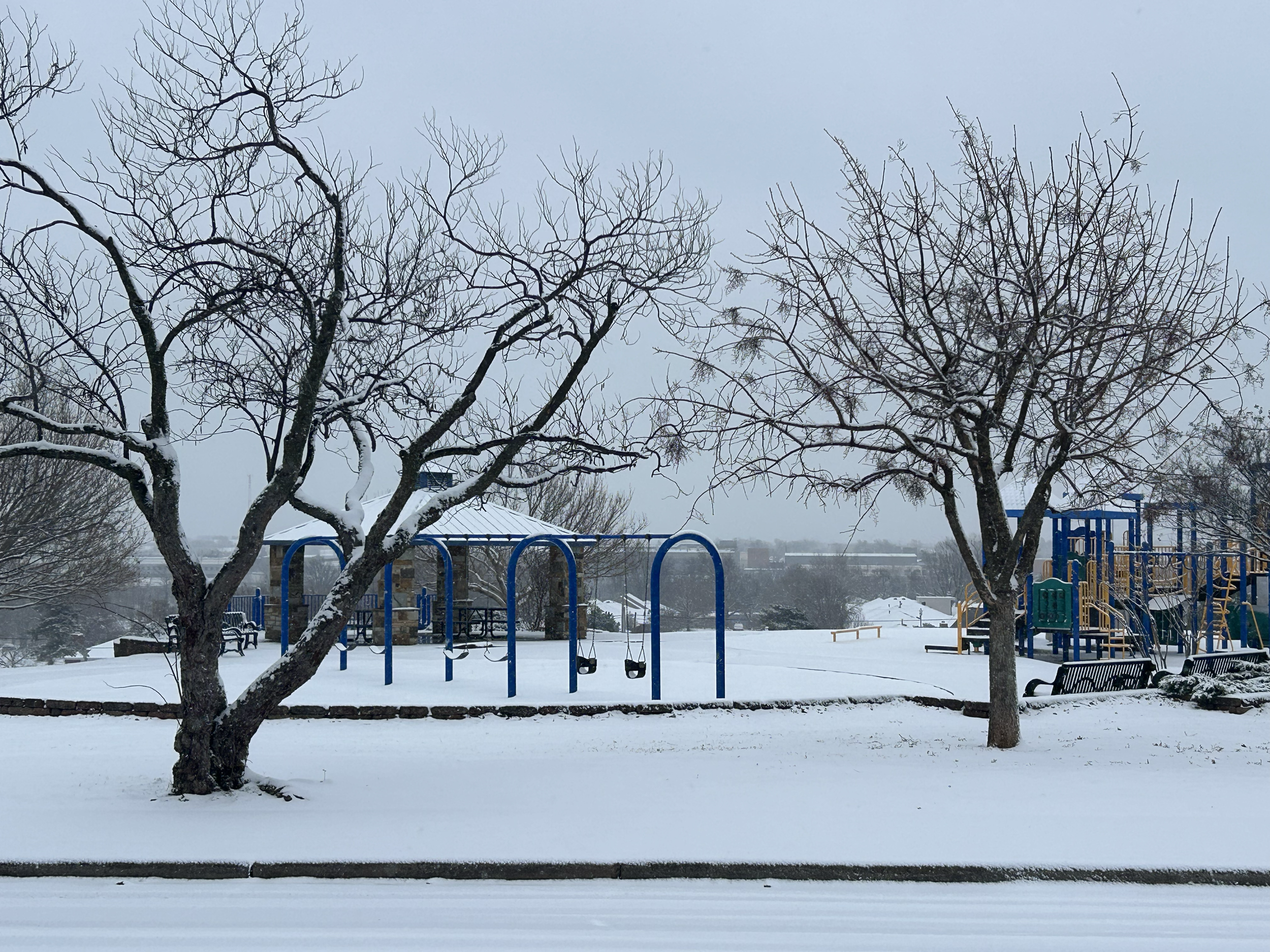 Snow-covered playground with two leafless trees, blue swing set, benches, and a small shelter, under a cloudy sky.
