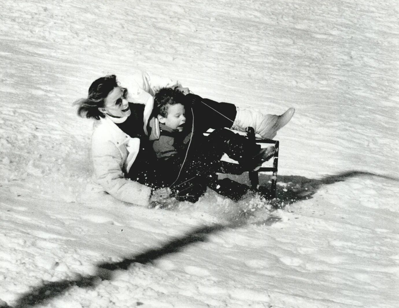 A woman and a young boy are riding down a snowy hill on a sled. The woman is smiling and wearing sunglasses, with her hand on the boy's back for support. The boy is bundled up in winter clothing, holding onto the sled, with snow flying around as they go down the hill.