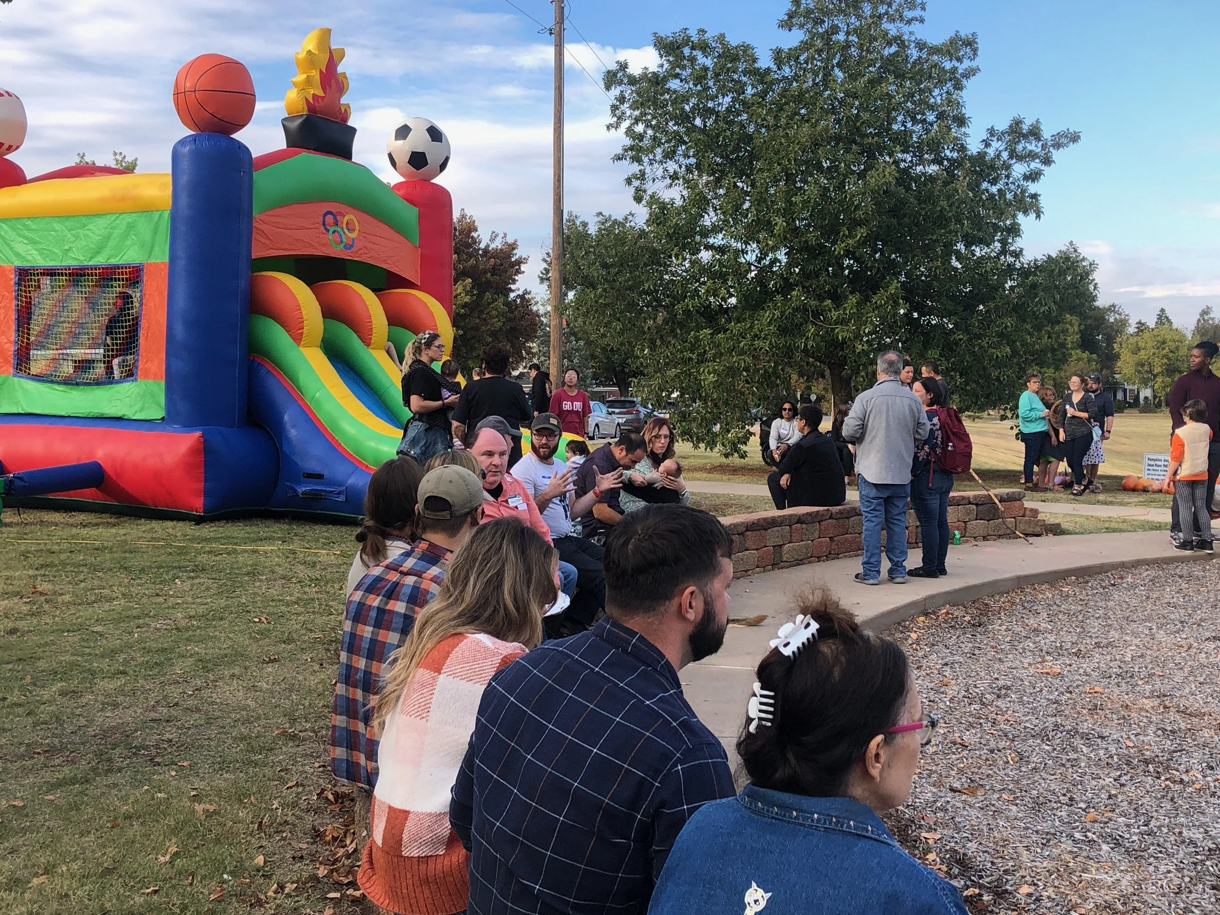 People gathered outdoors near a colorful inflatable bounce house with a slide, under a large tree, during a community event.