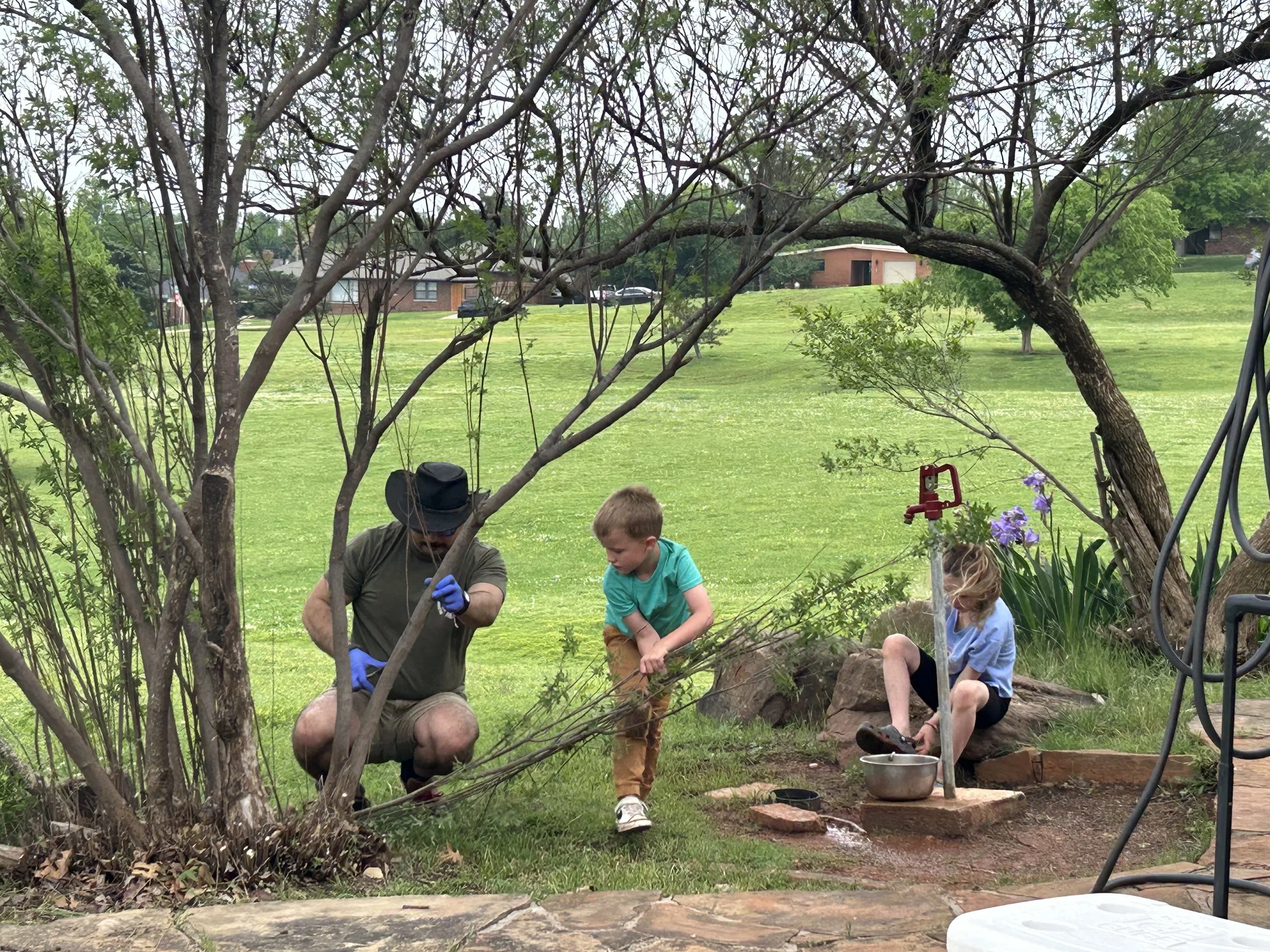 A man and two children working outdoors in a garden, planting or tending to a bush near a tree with houses in the background.