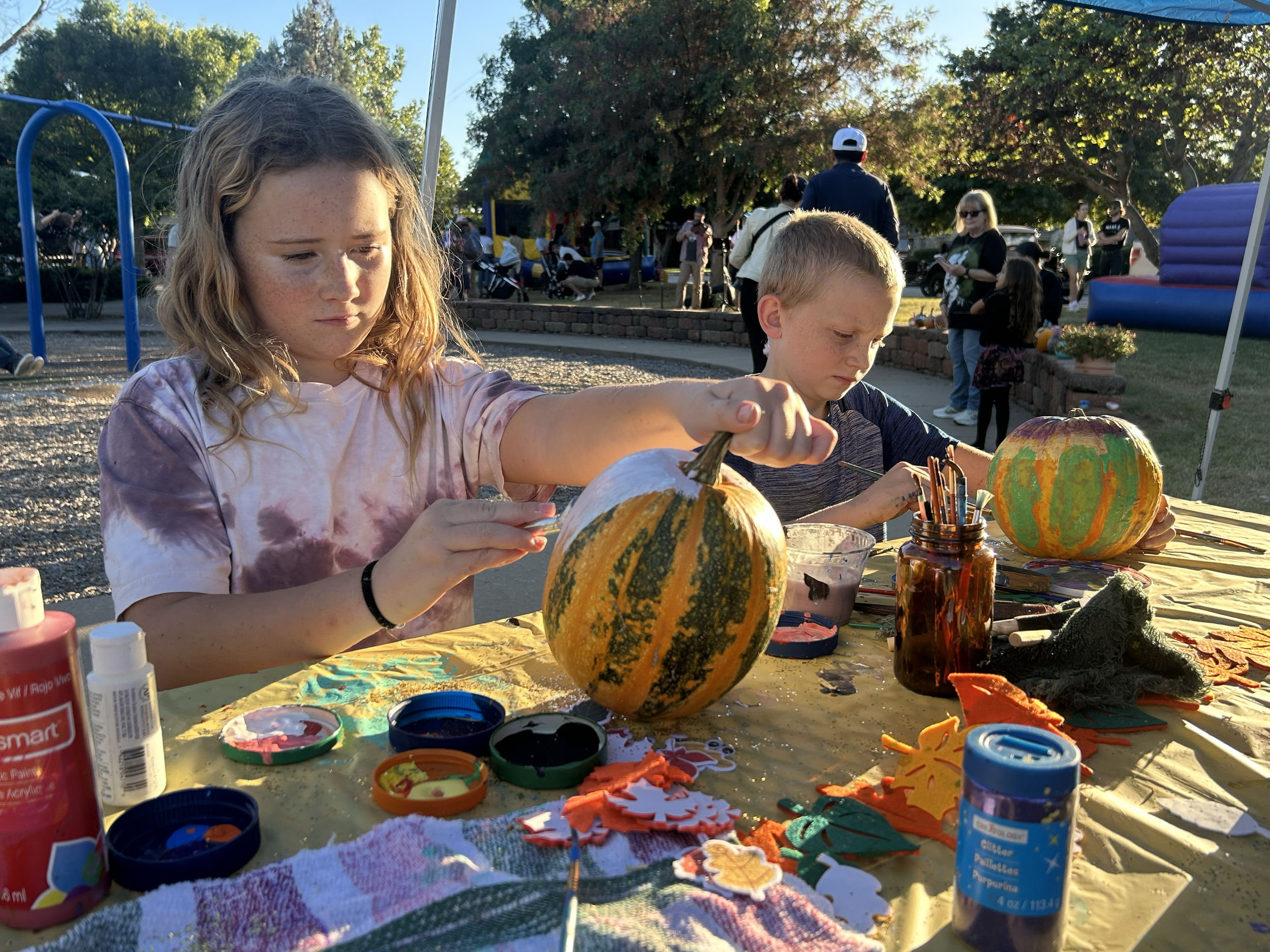 Two children decorating pumpkins at a craft table outdoors during daytime, with onlookers and inflatable bounce houses in the background.