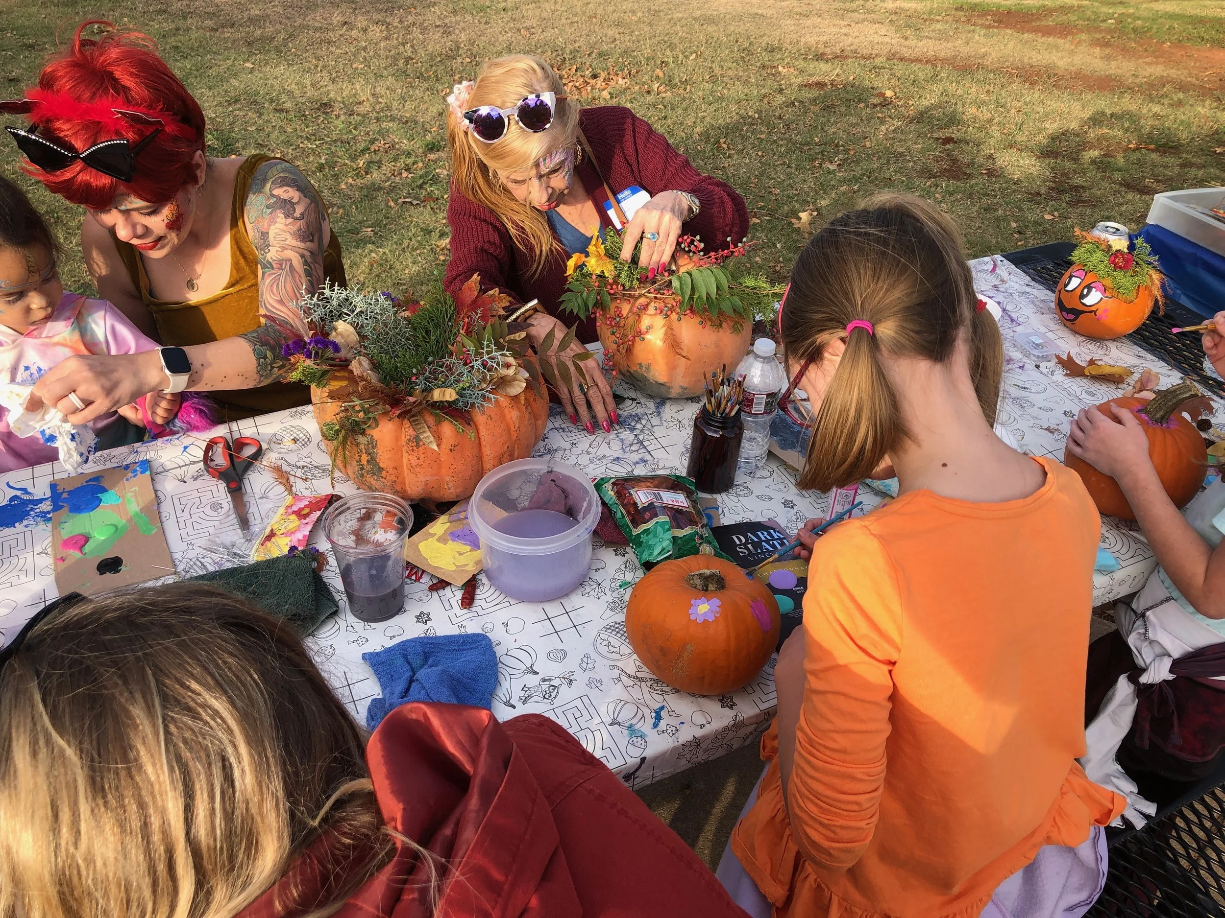 A group of children and women decorating pumpkins with flowers and paint at an outdoor gathering during fall.